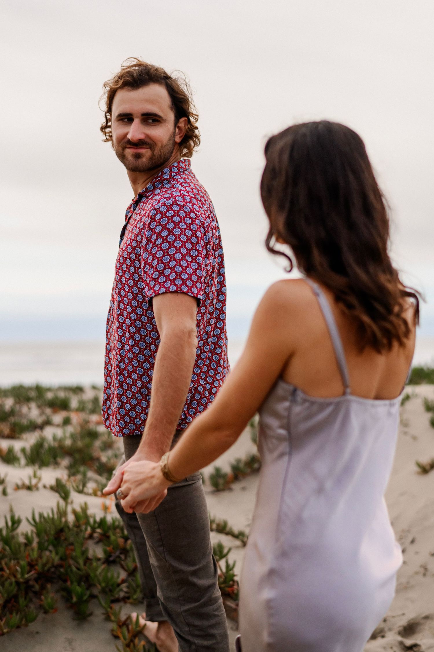 Curly-haired man in patterned shirt smiling back while holding womans hand.