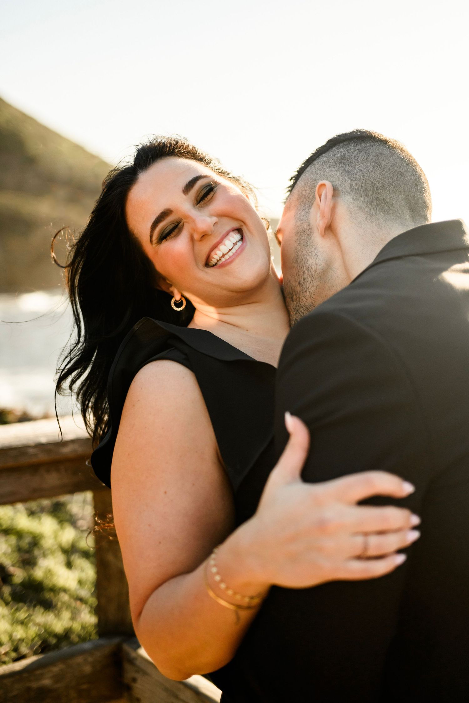 A joyful woman in a black dress laughs while embracing her partner near a coastal railing at golden hour.