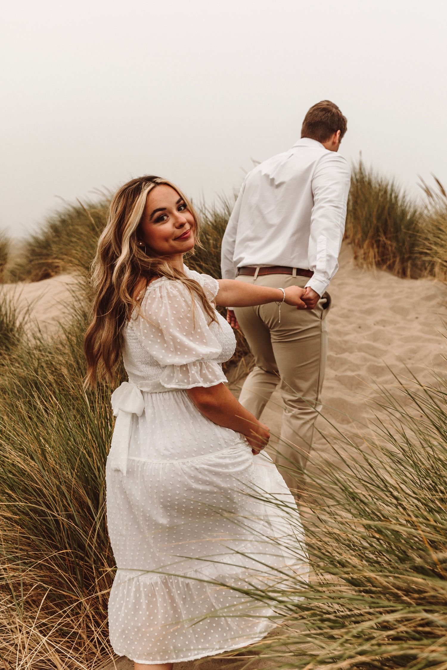 Blonde woman in white dress smiling at camera while holding hands with man on sandy dunes.