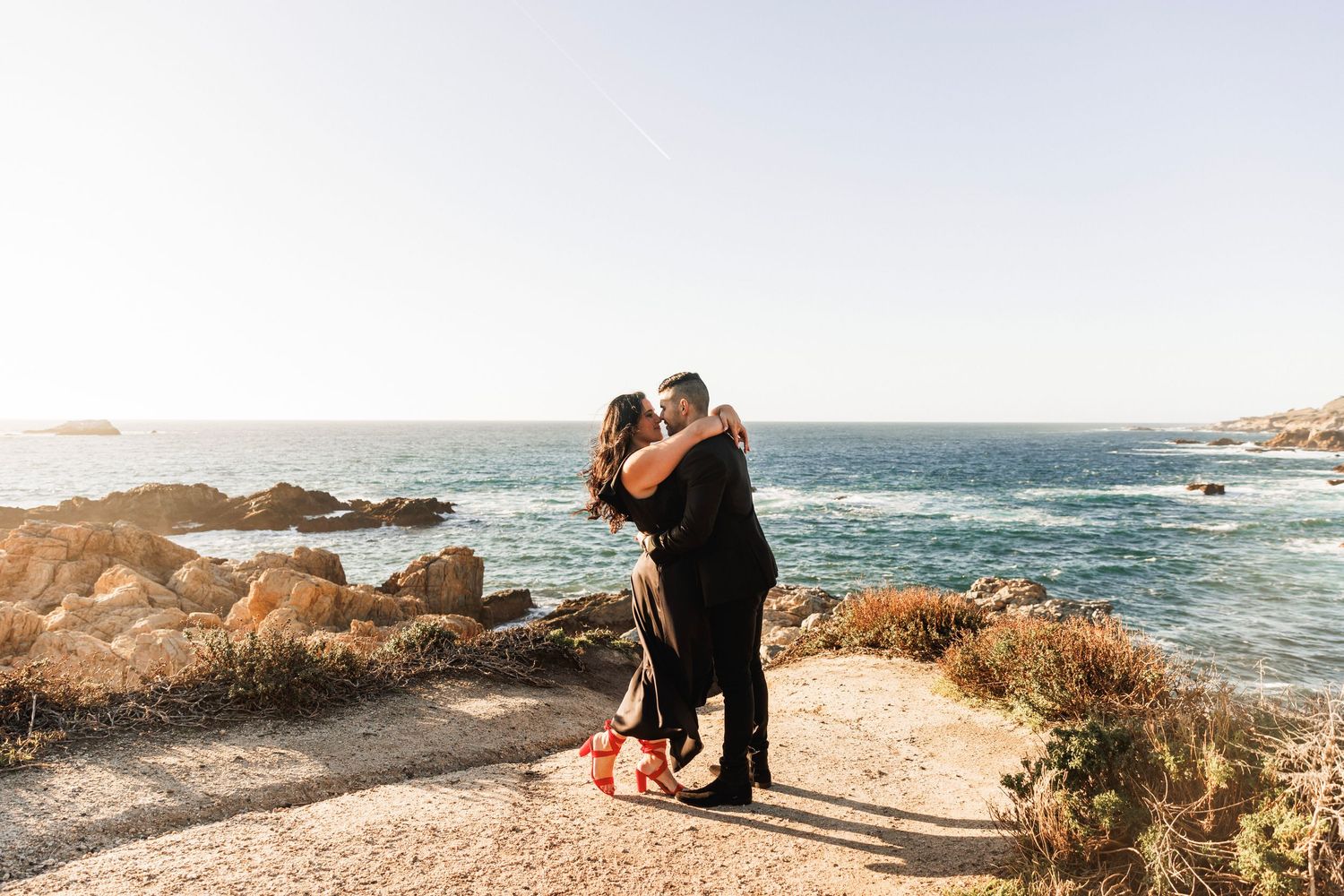 A couple shares a romantic embrace on rocky coastal cliffs with the blue ocean in the background.