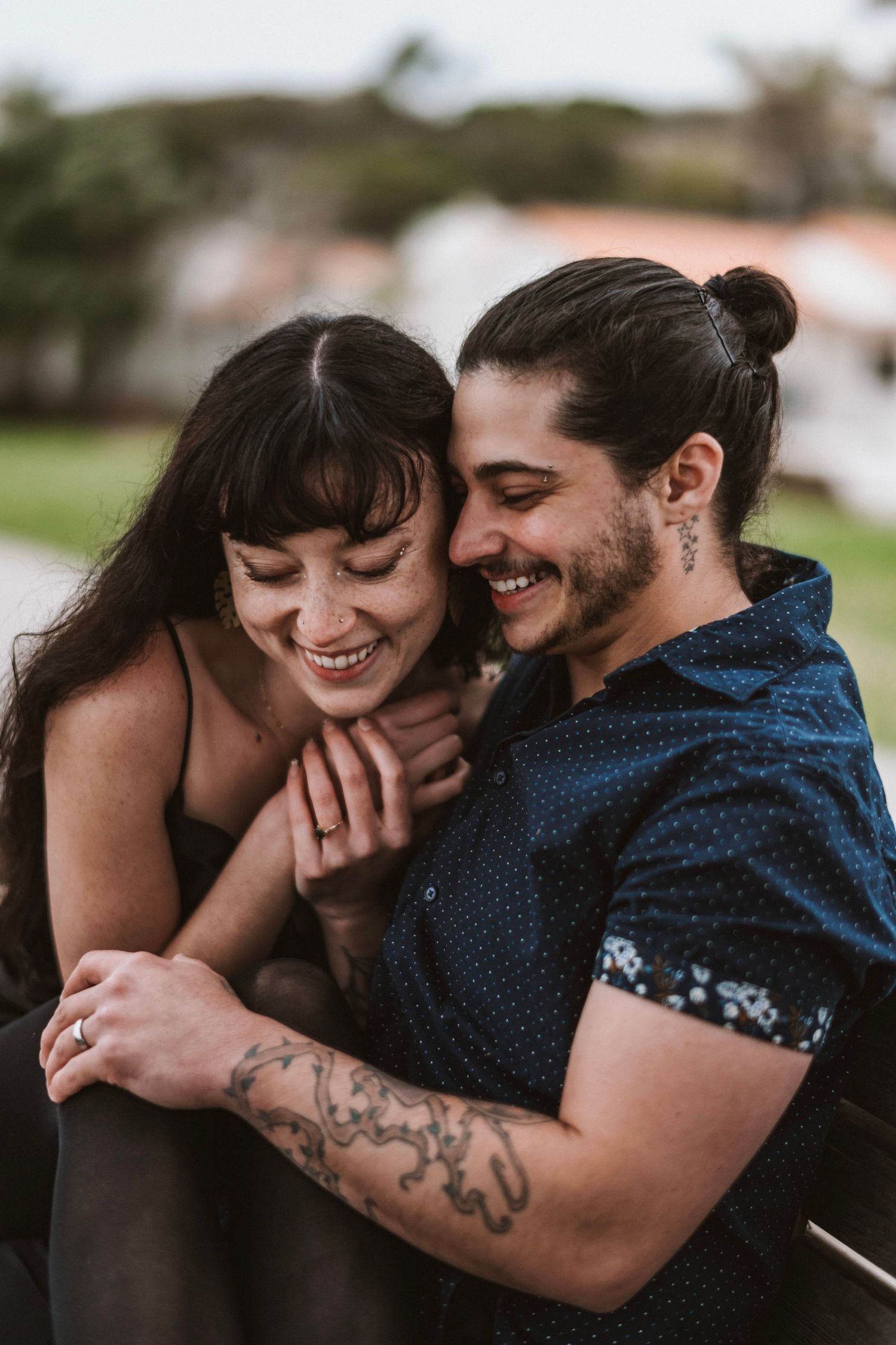 Happy couple embracing and laughing together outdoors, man with tattoos and bun hairstyle hugging smiling woman.