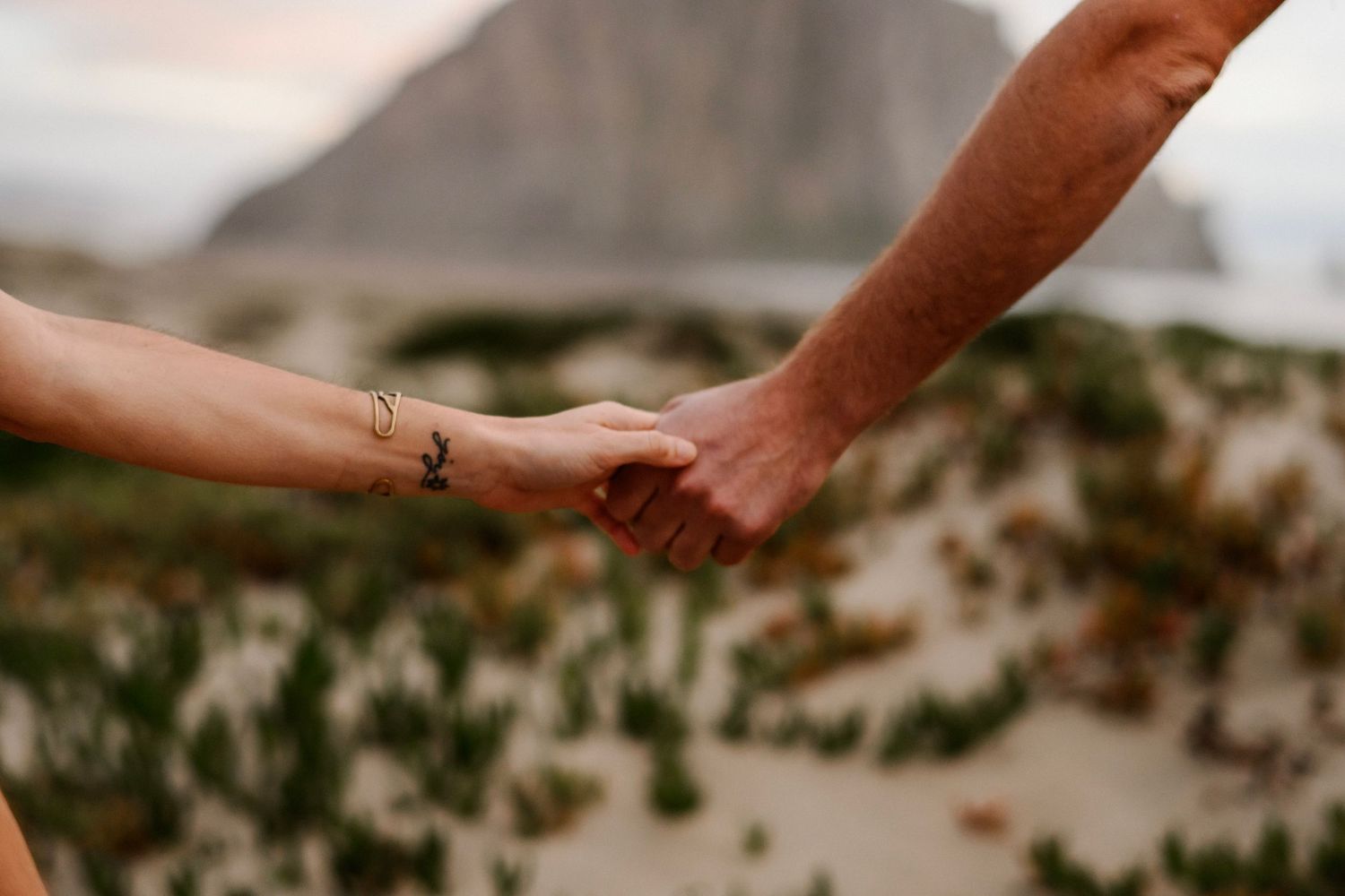 Close-up of couple holding hands on sandy beach with Morro Rock behind them.