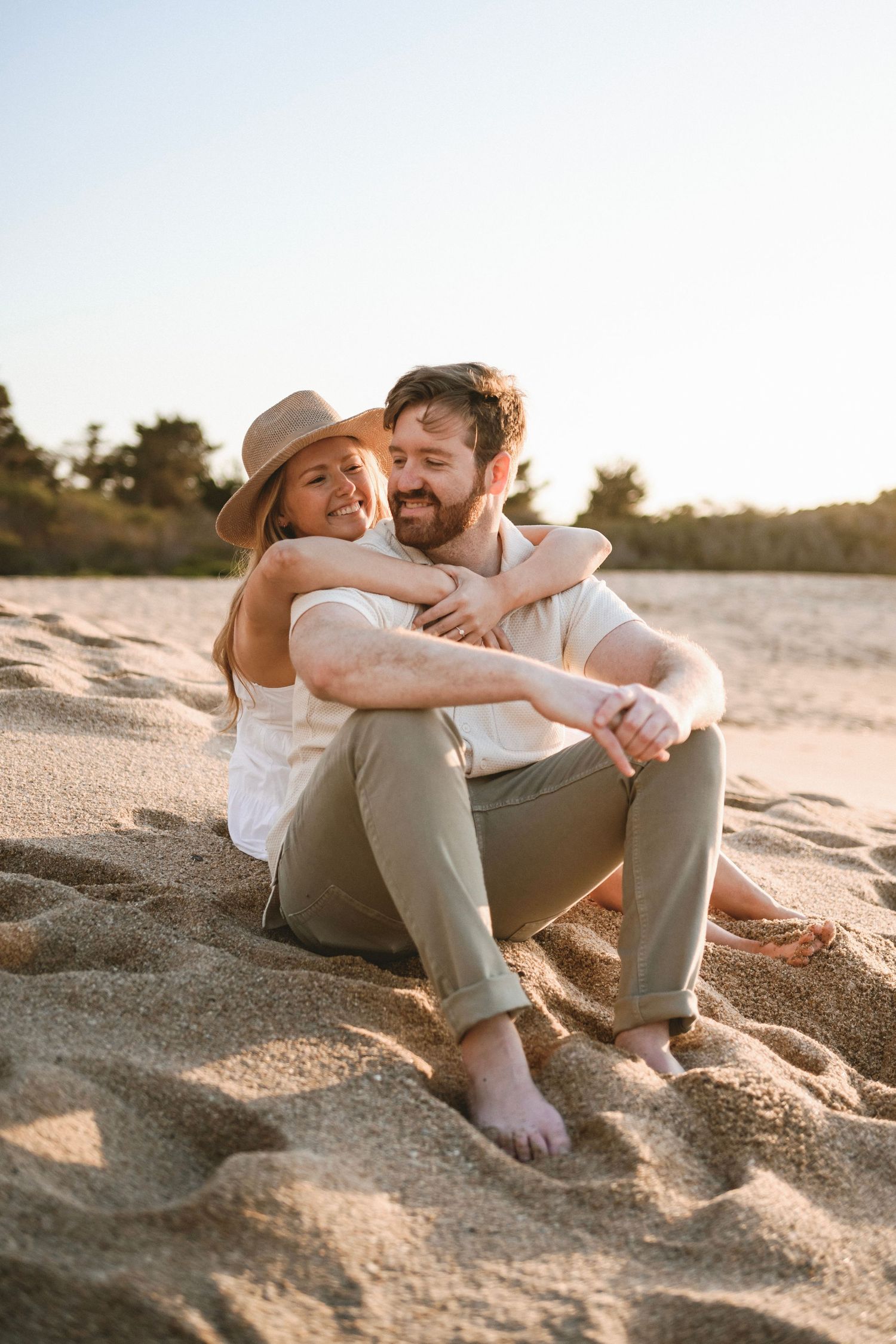 Couple sitting barefoot on sandy beach at sunset, embracing and smiling together.