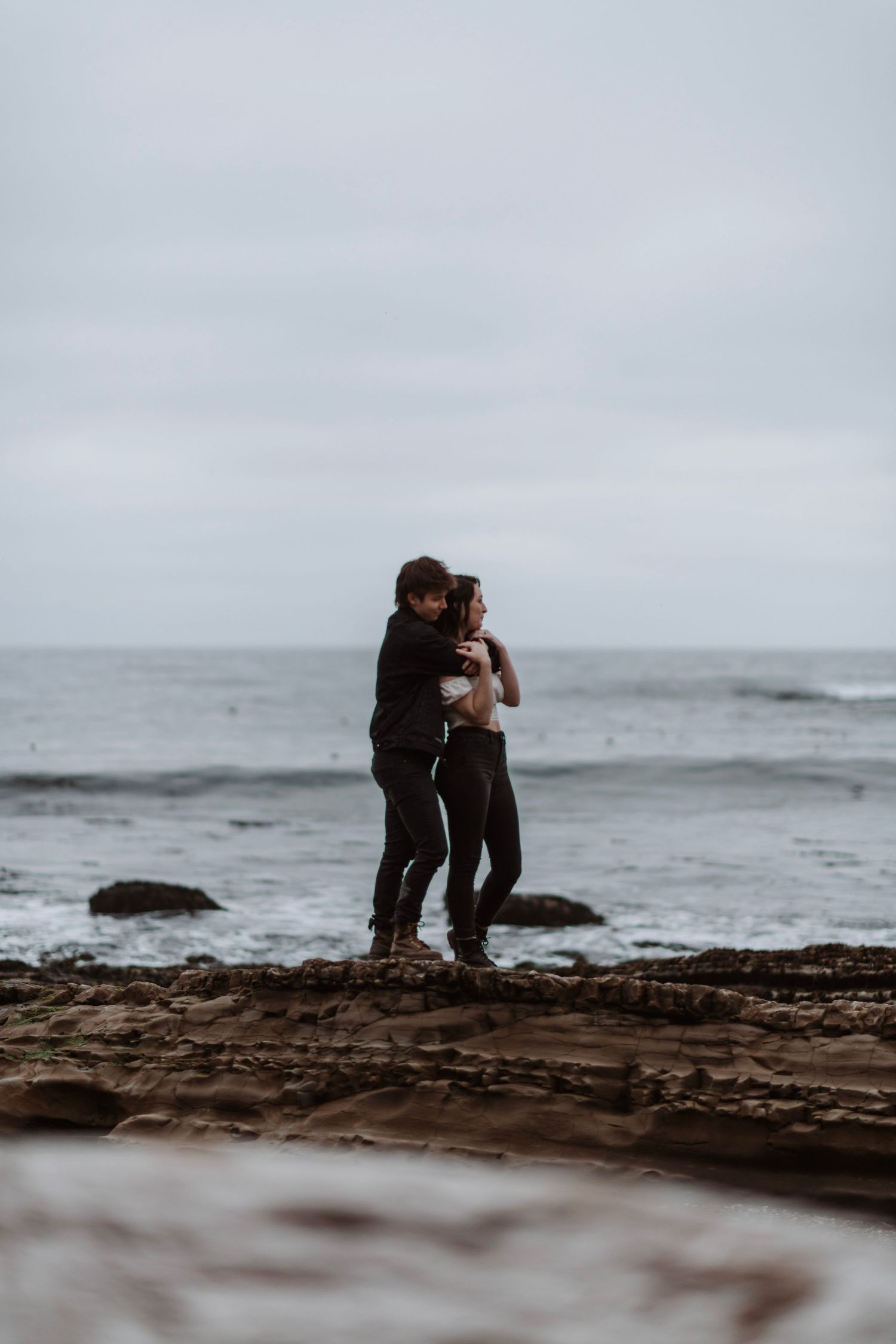Couple embracing on rocky shoreline with overcast ocean backdrop.
