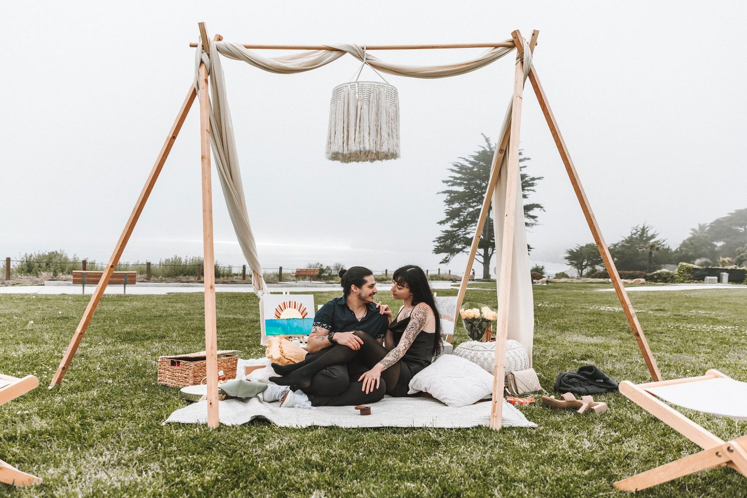 A couple enjoys a romantic boho-styled picnic setup on a lawn under a wooden arch with draping.