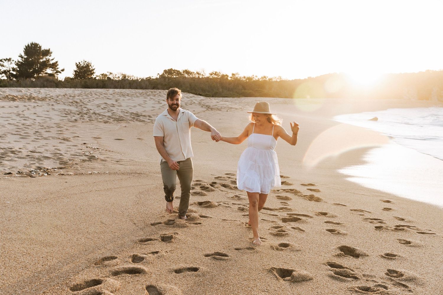 Happy couple running barefoot on golden sandy beach at sunset, woman in white dress holding hands.