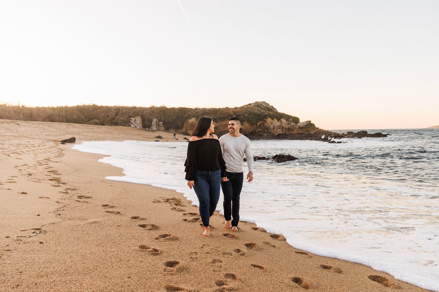 Couple walking barefoot along ocean shoreline at sunset, leaving footprints in wet sand.
