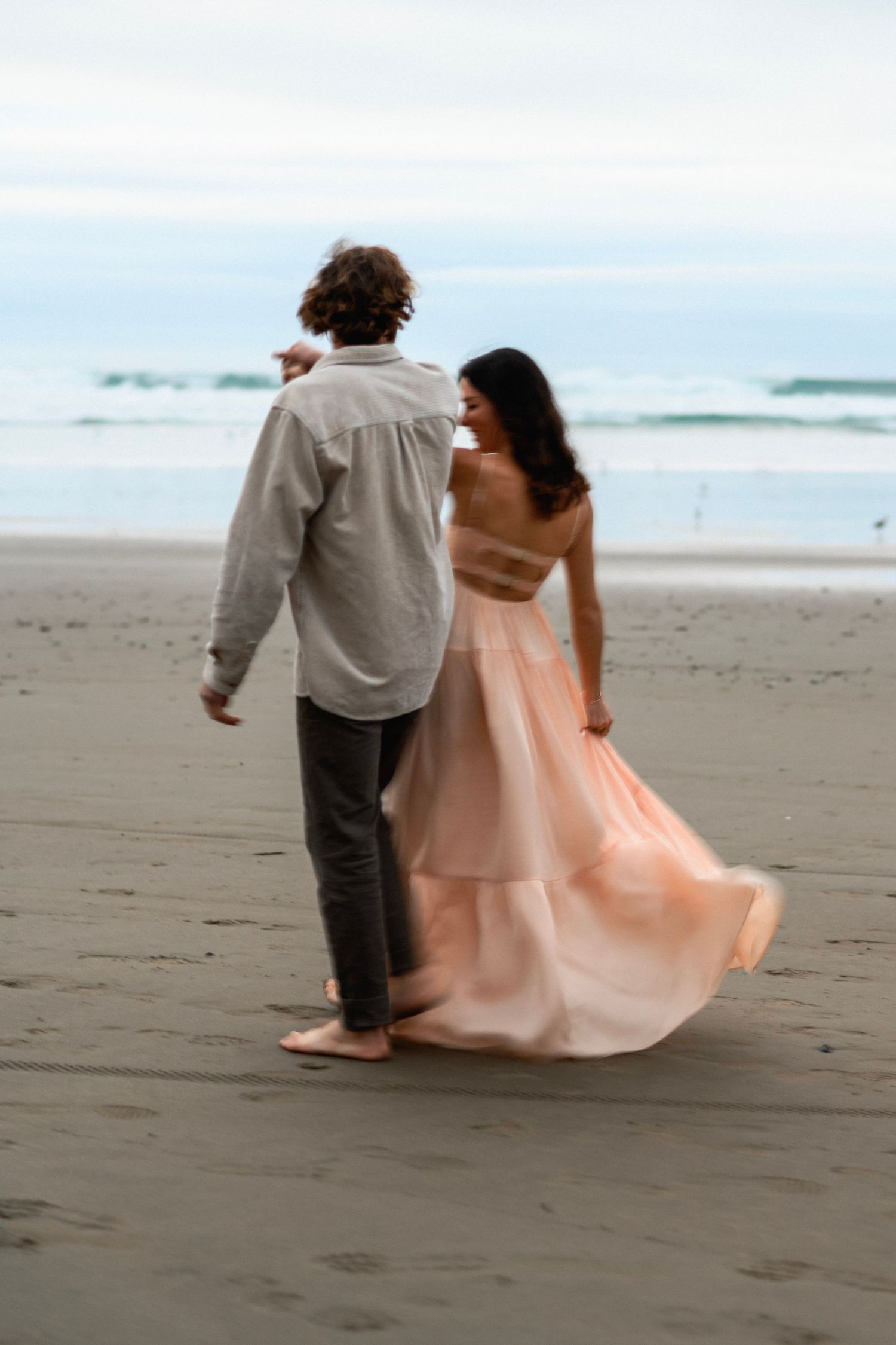 Couple walking on beach, woman in flowing pink gown, man in gray shirt, ocean waves in background.