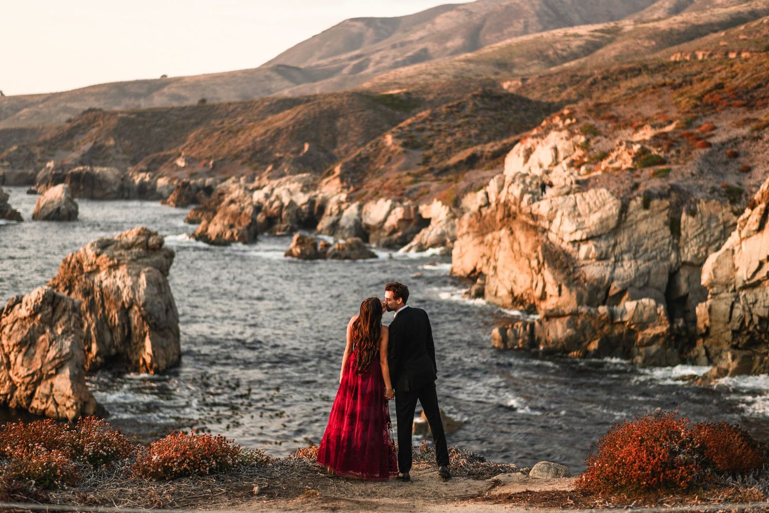 Couple sharing a tender moment on a coastal cliff overlooking rocky ocean shoreline, woman in red gown.