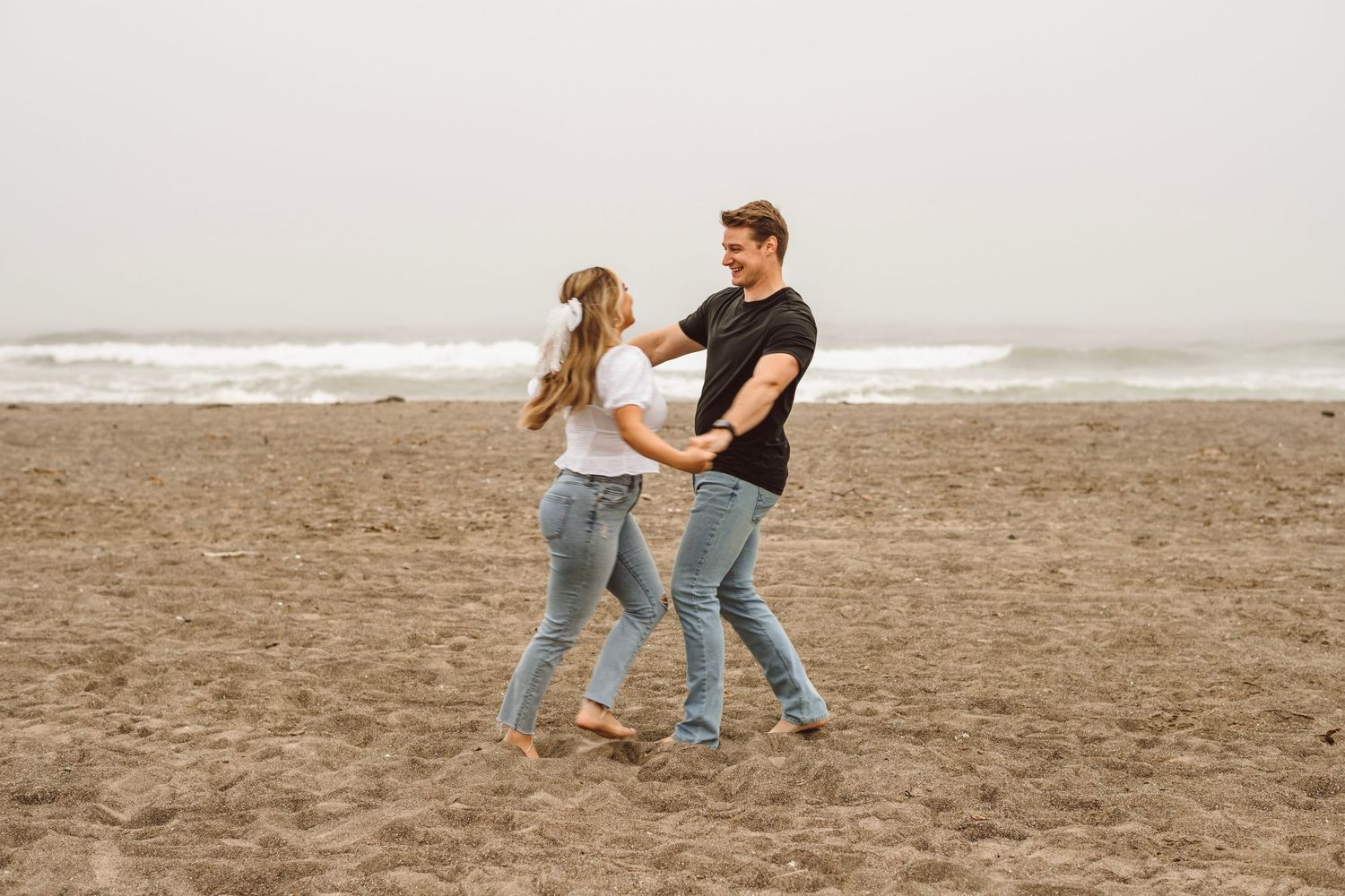 Carefree couple dancing and laughing together barefoot on foggy ocean beach in casual outfits.