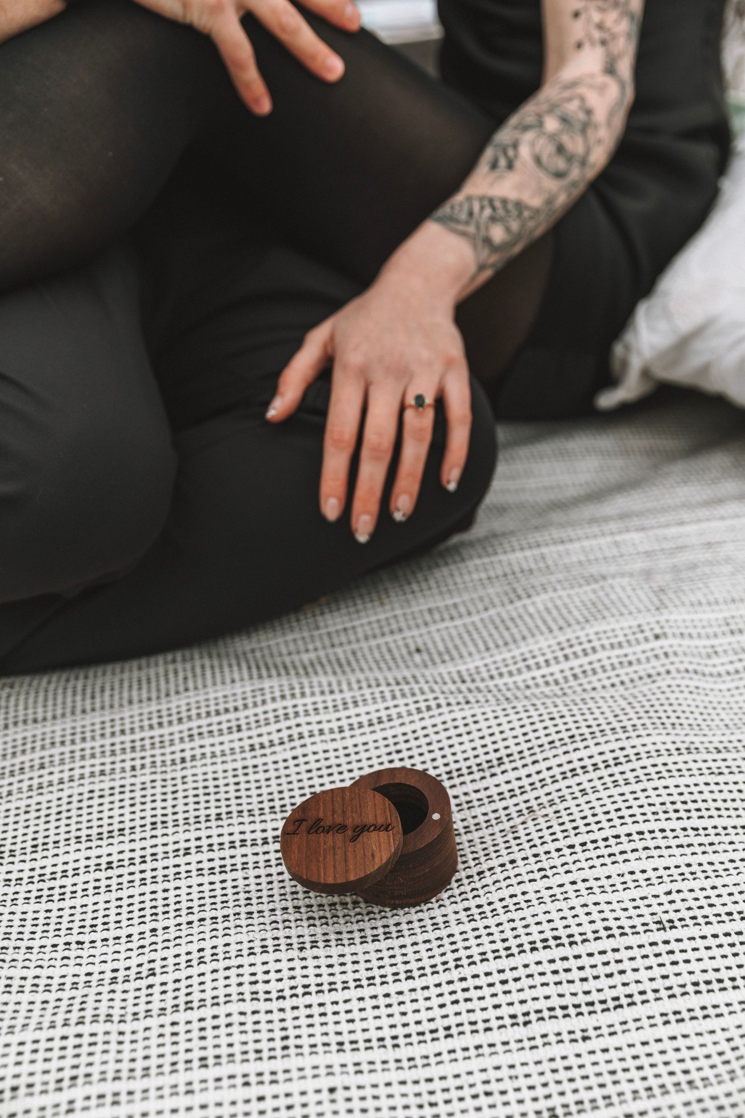 A close-up of a tattooed hand wearing a dark engagement ring beside a wooden ring box on a blanket.