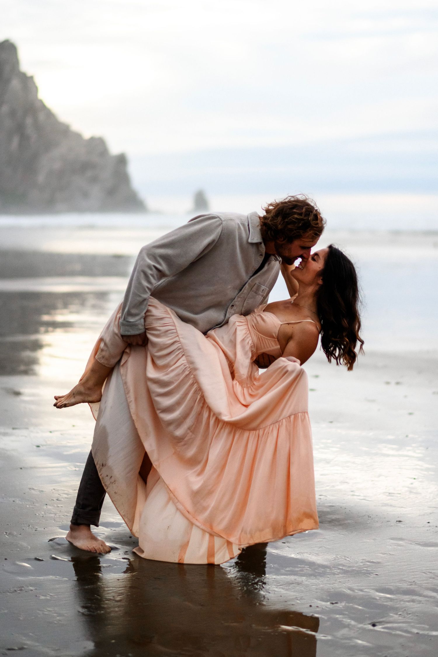 Couple sharing a passionate dip kiss on a reflective wet beach with dramatic rocky cliffs in background.