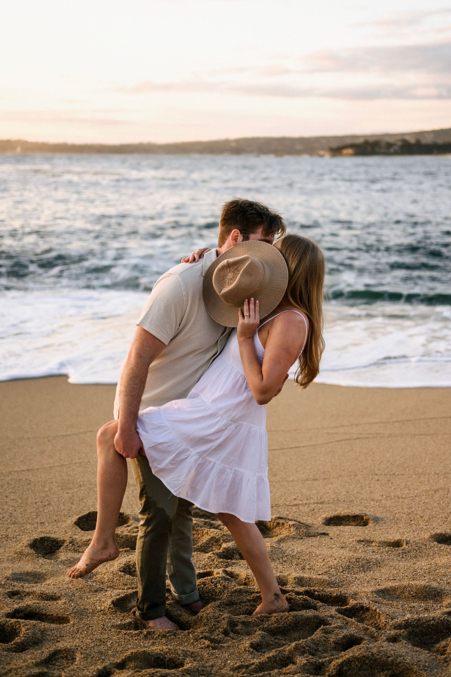 Couple sharing a kiss on a sandy beach at sunset with ocean waves behind them.