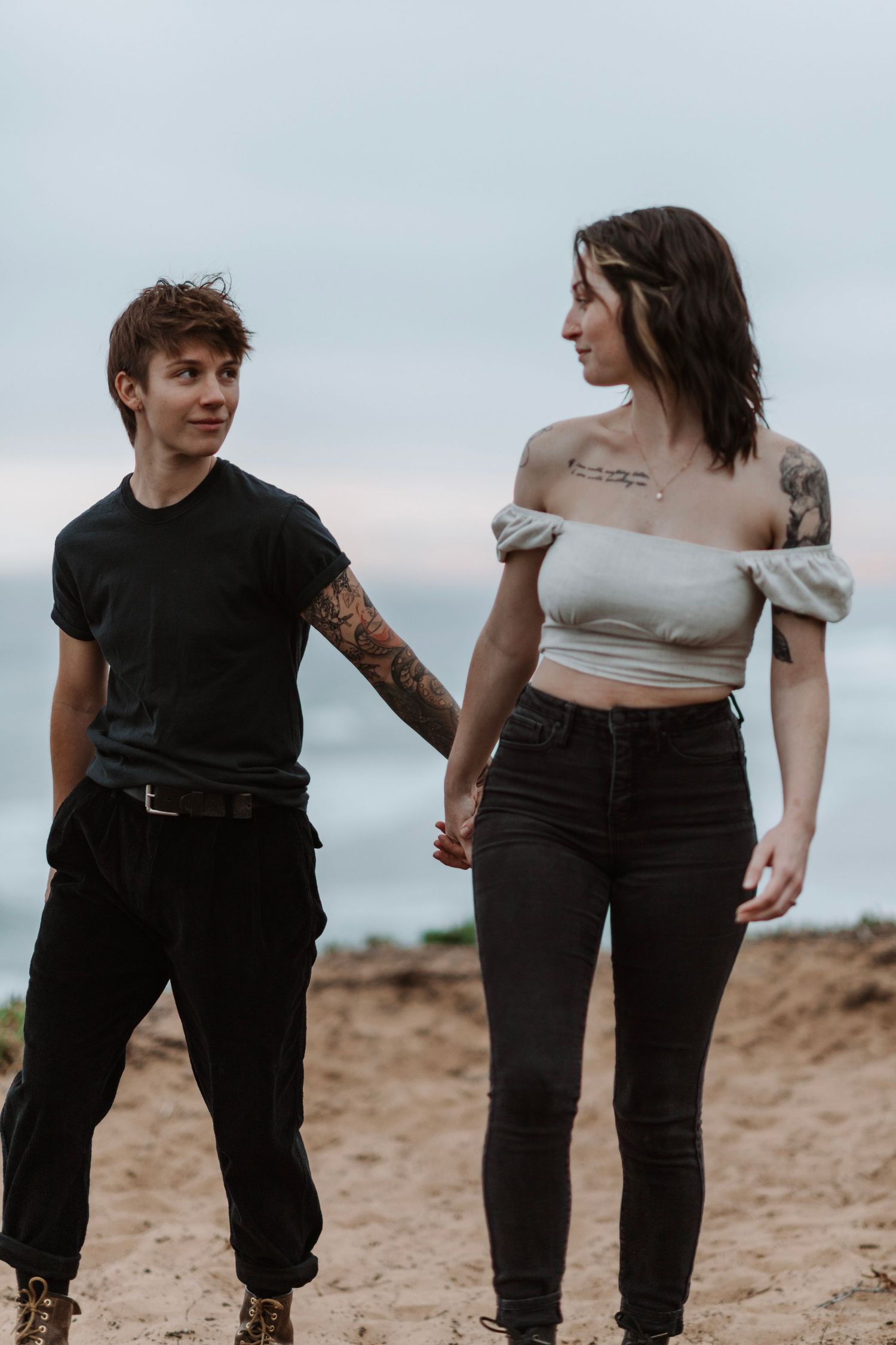Tattooed couple holding hands and smiling at each other on sandy cliffs.