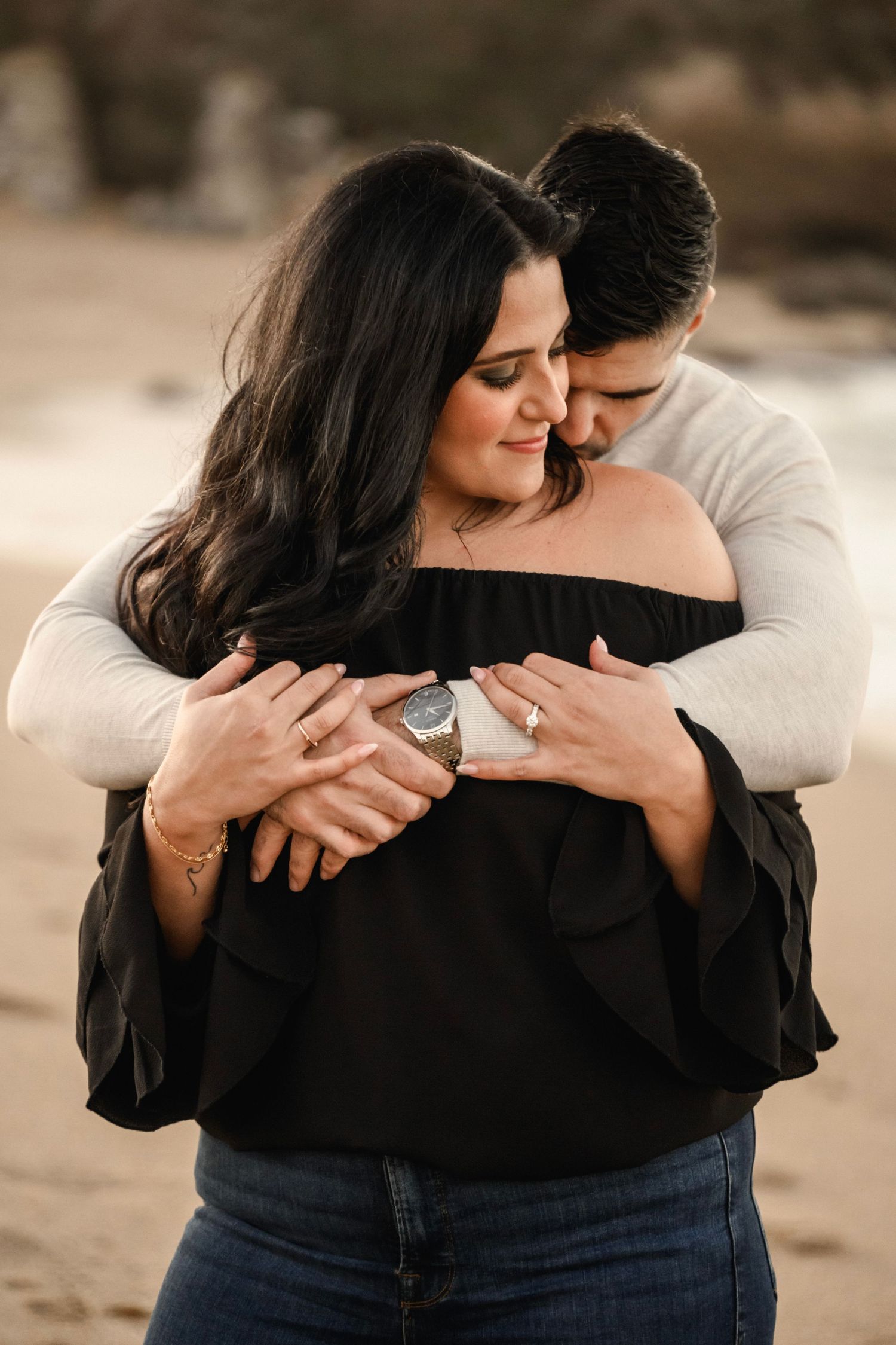 A man tenderly hugs a woman in an off-shoulder black top from behind on a sandy beach.