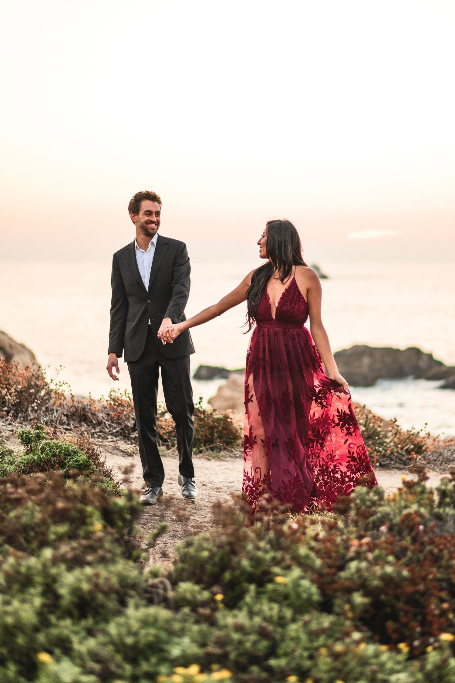 A couple walks hand in hand along a coastal path at sunset, the woman in a flowing red dress.