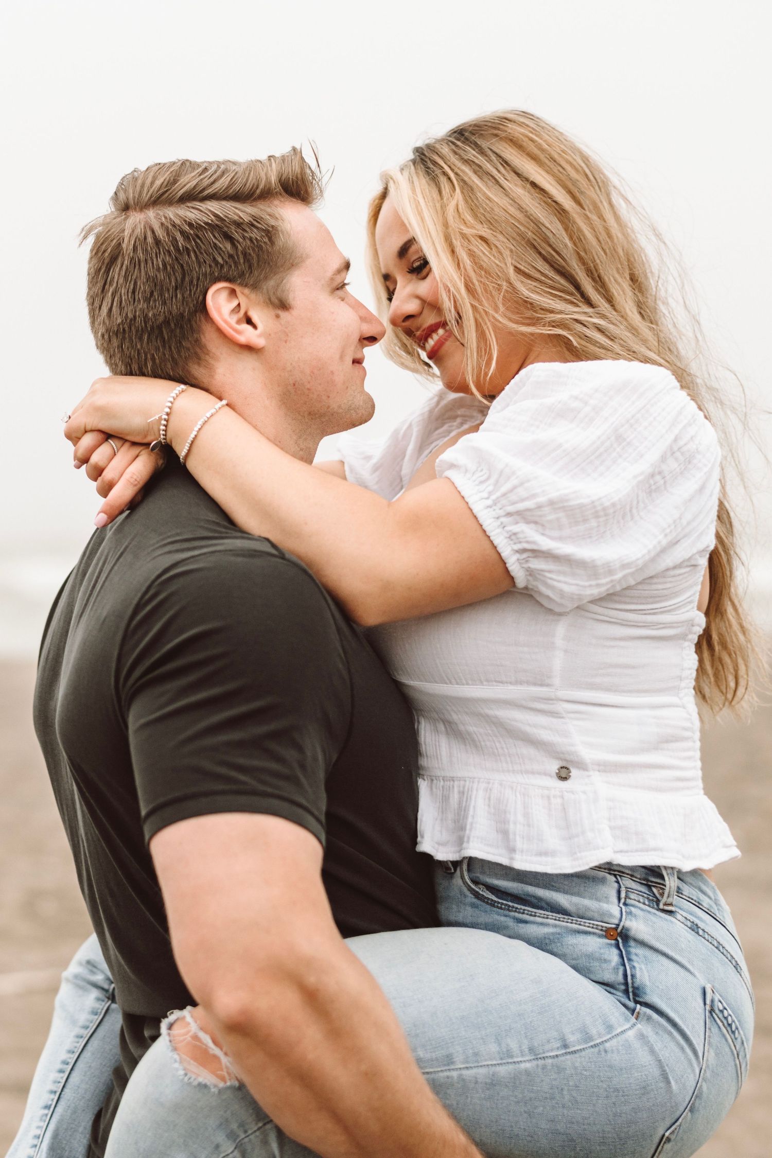 Man lifting smiling blonde woman on foggy beach, both laughing in casual beach attire.