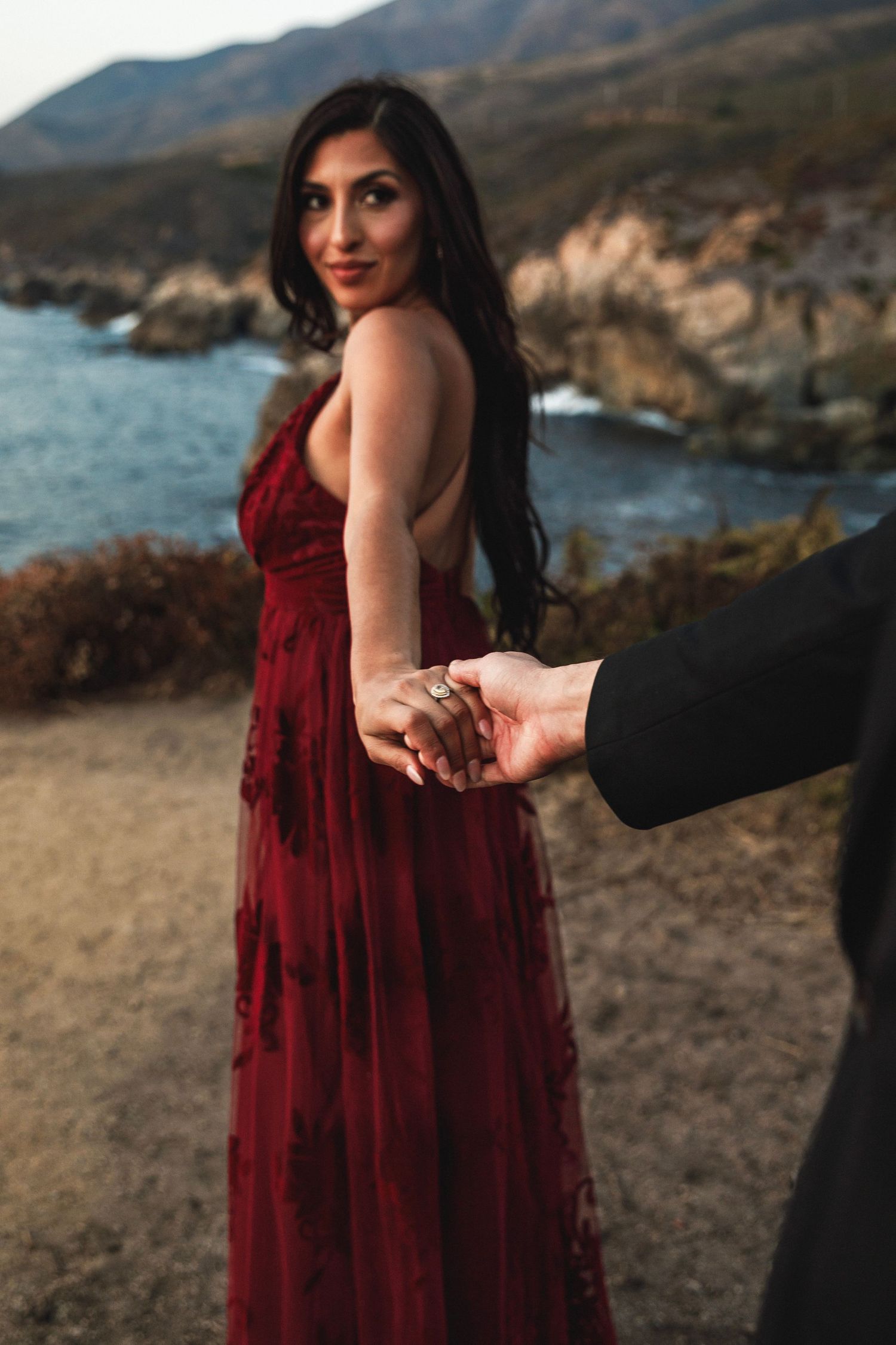A woman in a stunning backless burgundy gown holds her partners hand near a scenic coastal waterway.