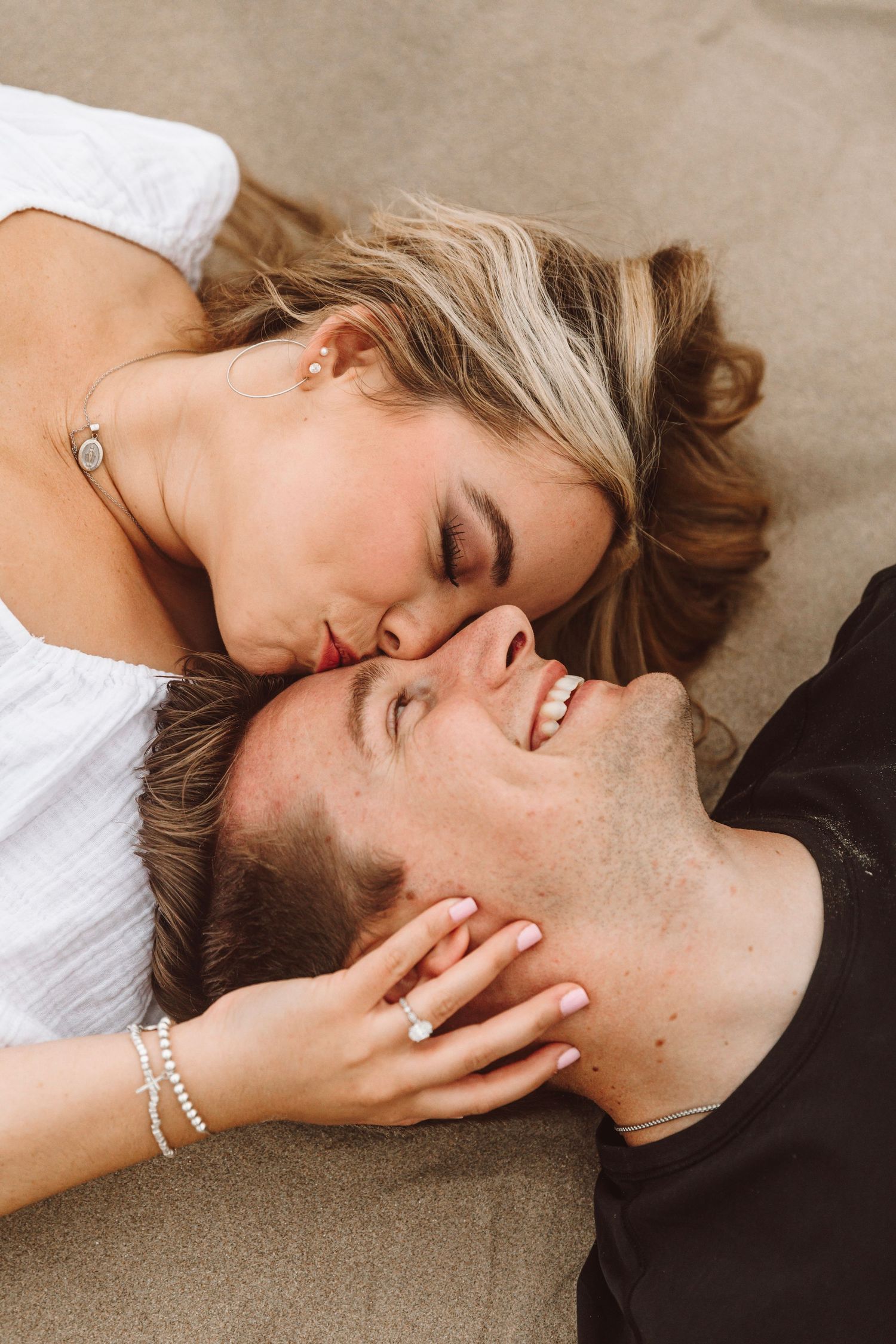 Engaged couple lying on sand sharing a kiss with diamond engagement ring visible on her hand.