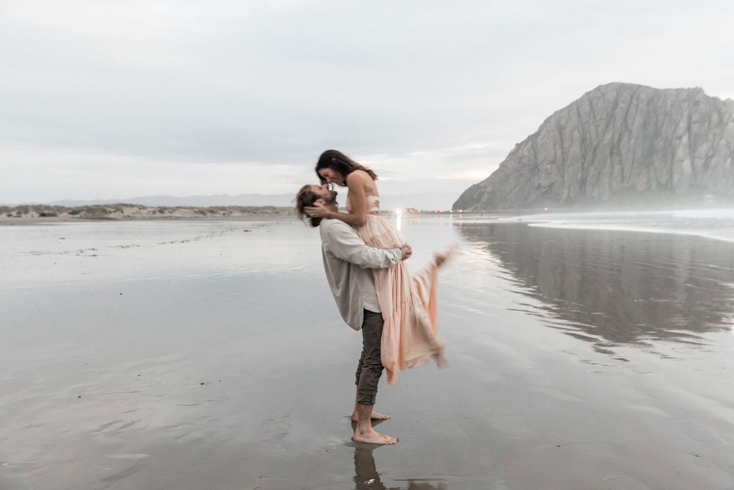 Romantic couple embracing on wet beach with dramatic rock formation reflected in water behind them.