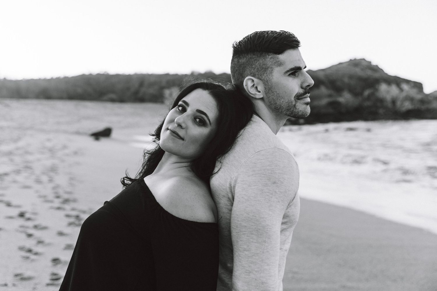 A black and white photo of a couple standing back to back on a serene beach shoreline.