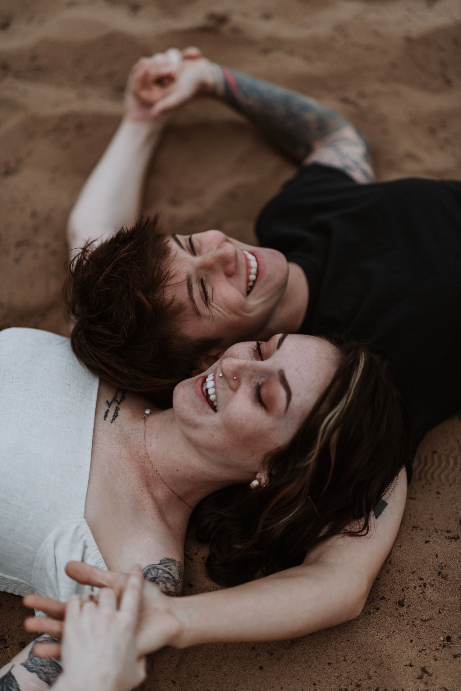 Joyful tattooed couple lying on sandy beach laughing together, holding hands overhead in a candid moment.