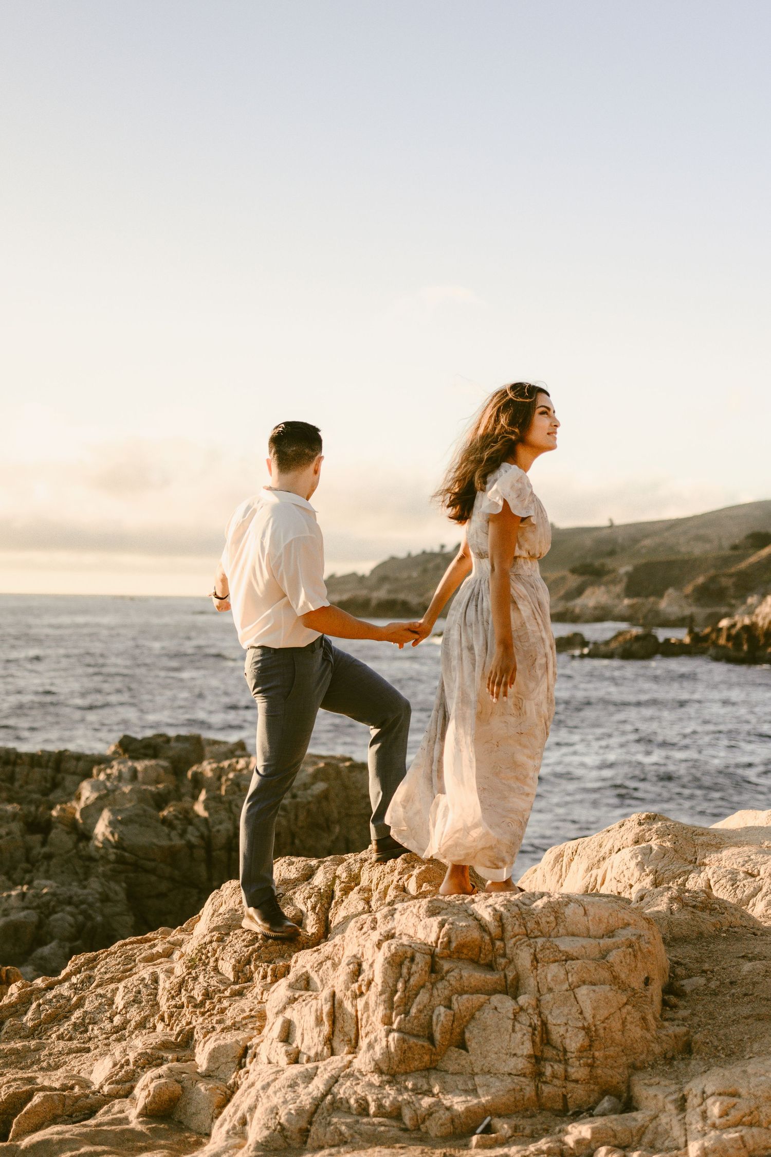 Couple holding hands standing on coastal rocks at sunset with ocean waves behind them.