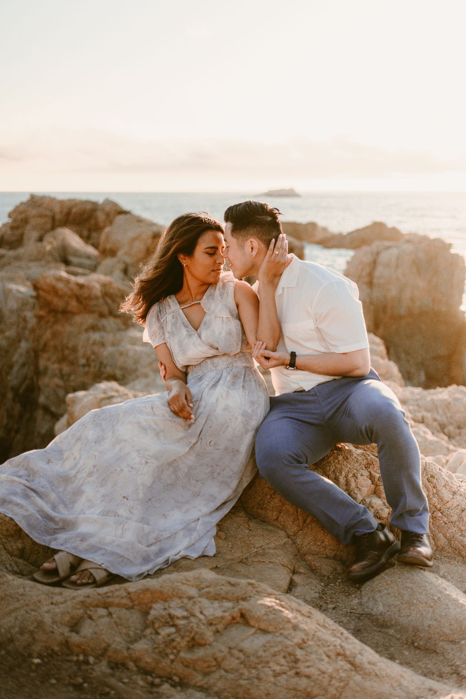Couple sharing a romantic moment sitting on coastal rocks during golden hour sunset.