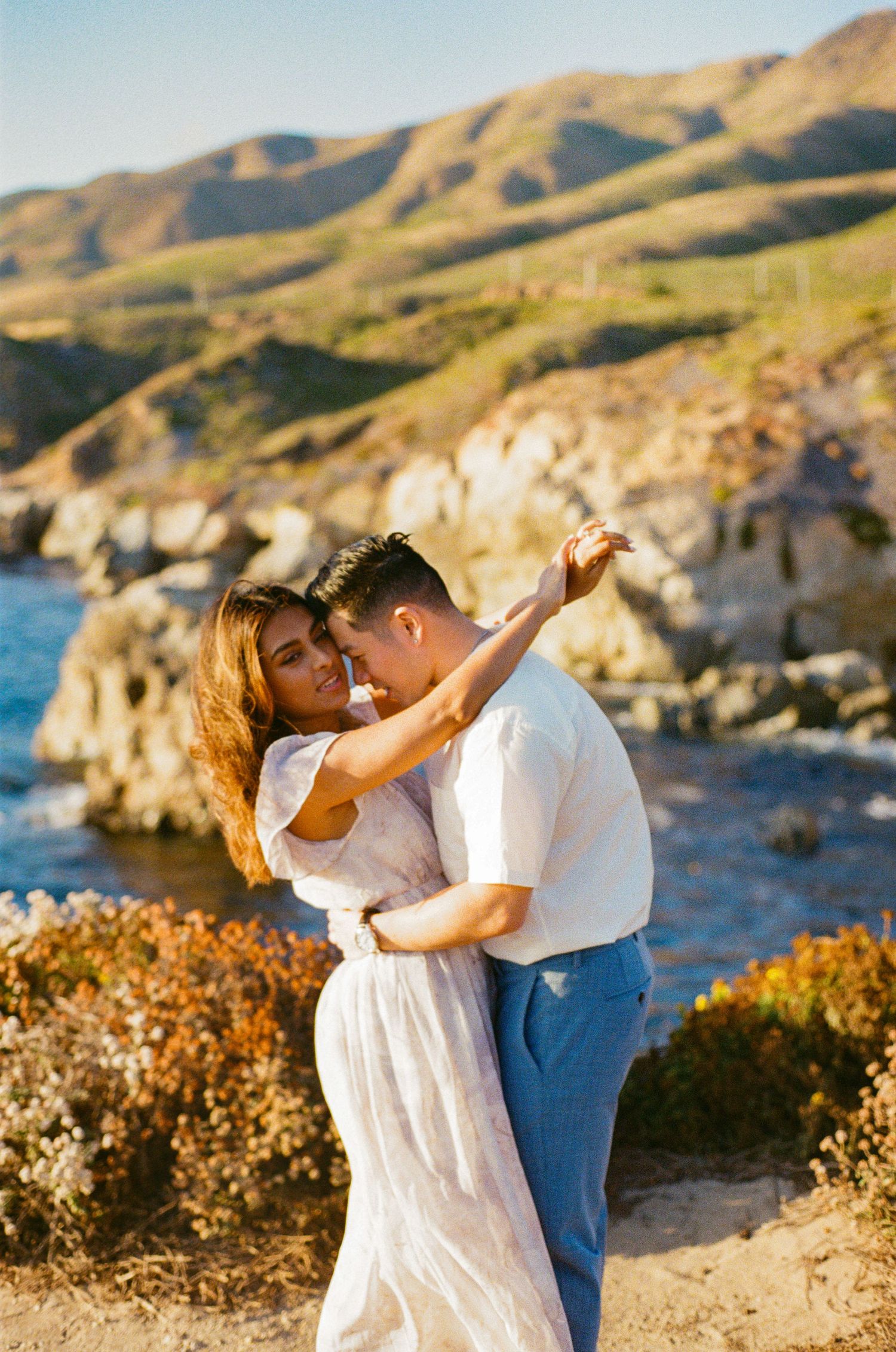 Couple embracing and dancing on a rocky California coastline during golden hour sunset.