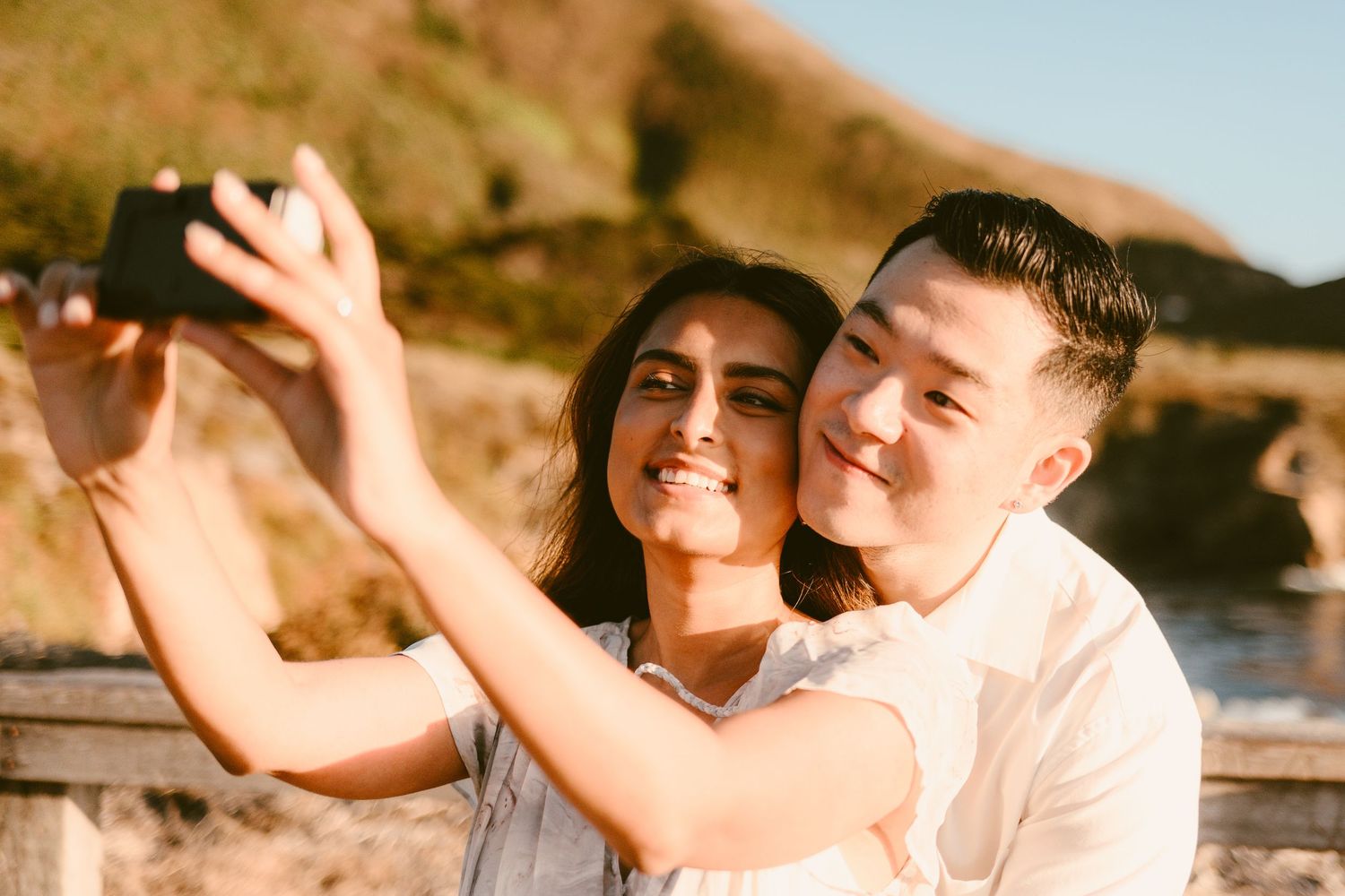 Smiling couple taking a selfie together with scenic coastal mountains in the background.