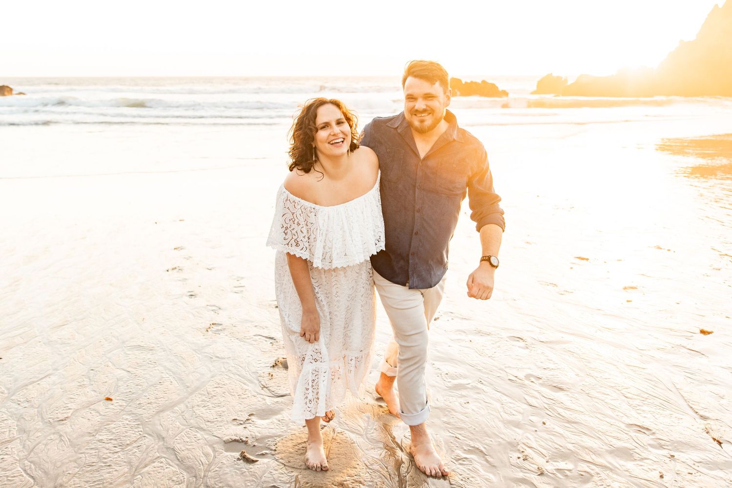Couple smiling and walking on a sunny beach at golden hour, woman in white dress, man in navy shirt.