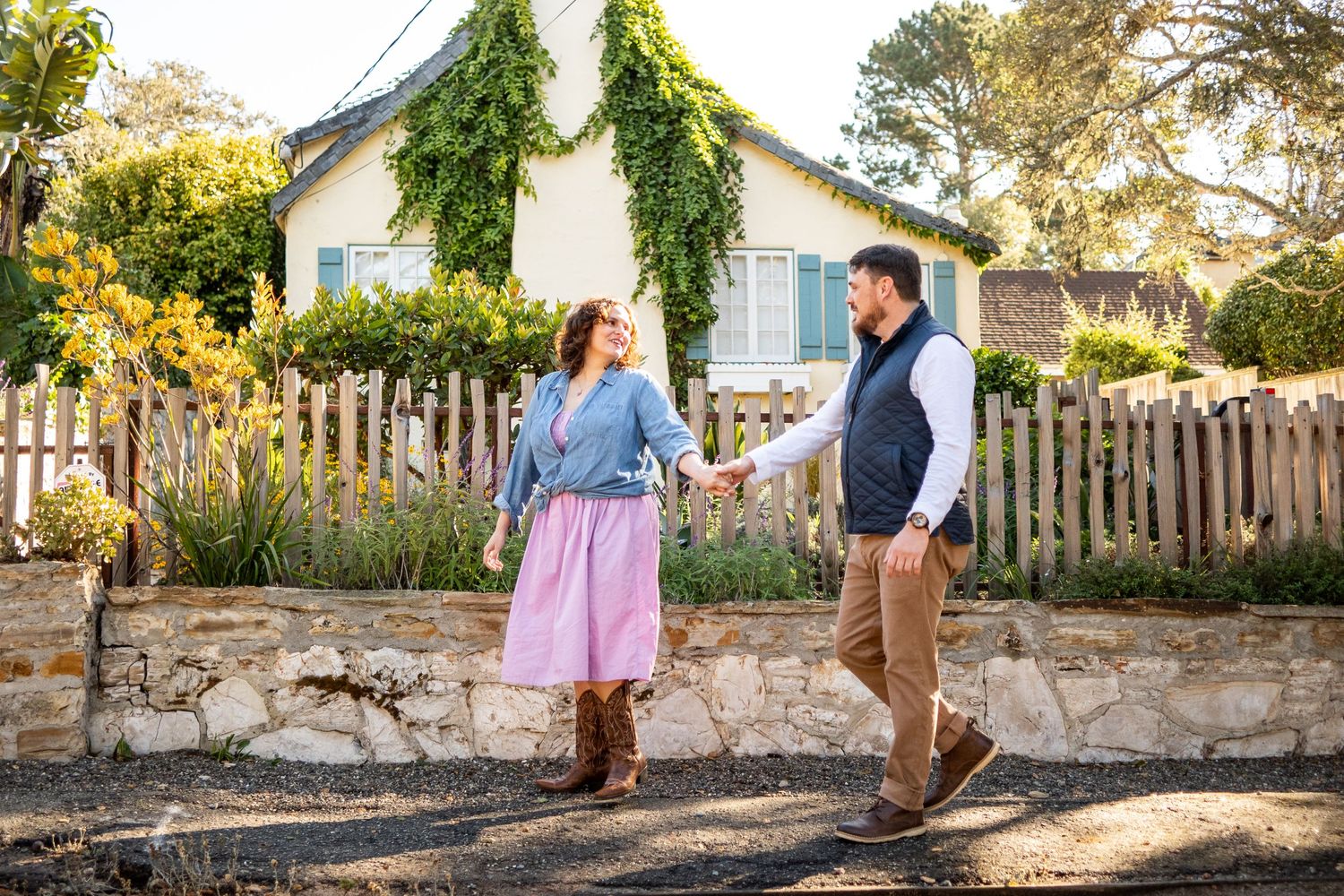 Couple holding hands in front of a charming cottage with ivy, woman in pink skirt and boots.