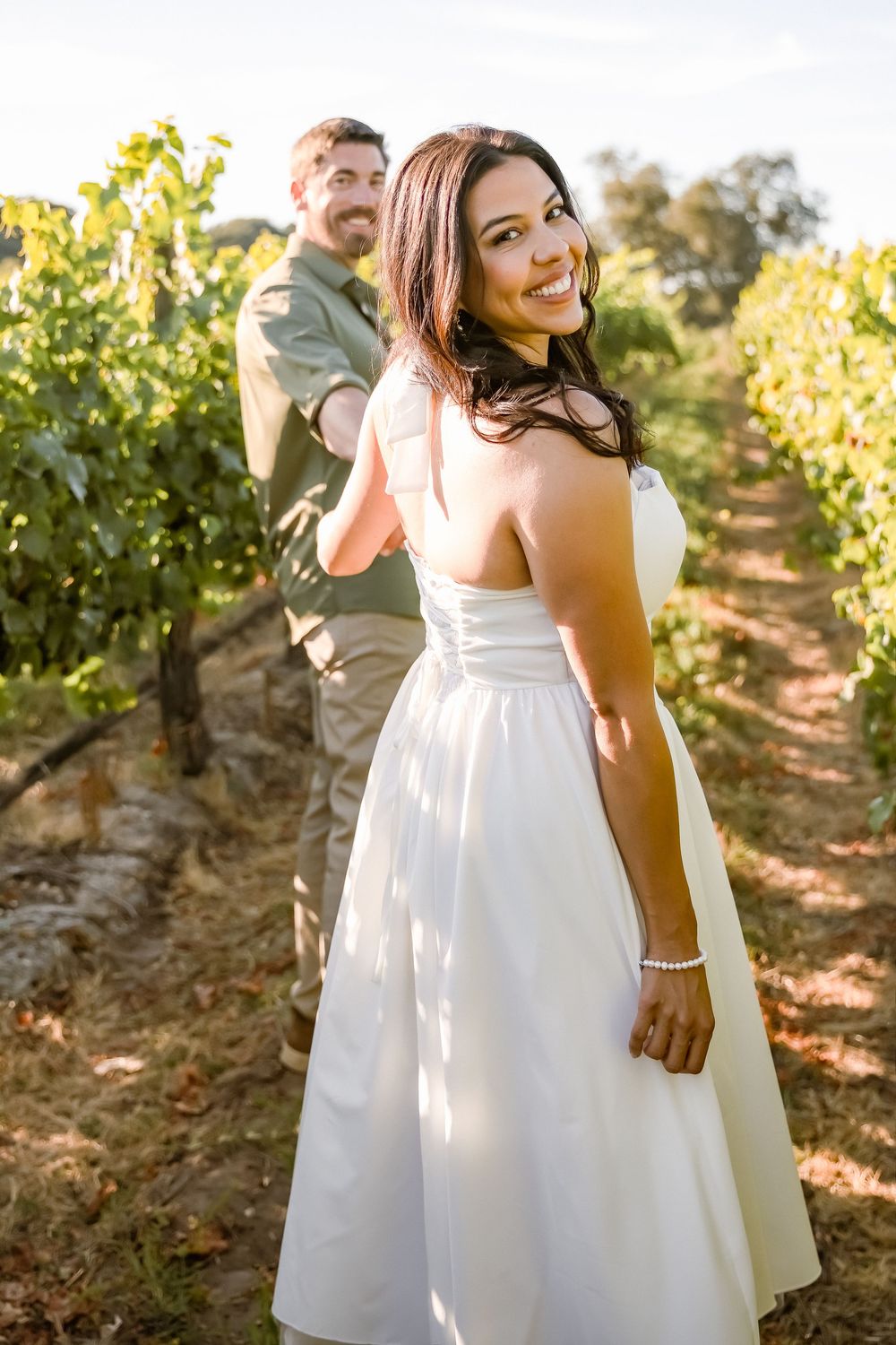 Bride in white dress smiles over shoulder holding grooms hand in sunny vineyard wedding photo.