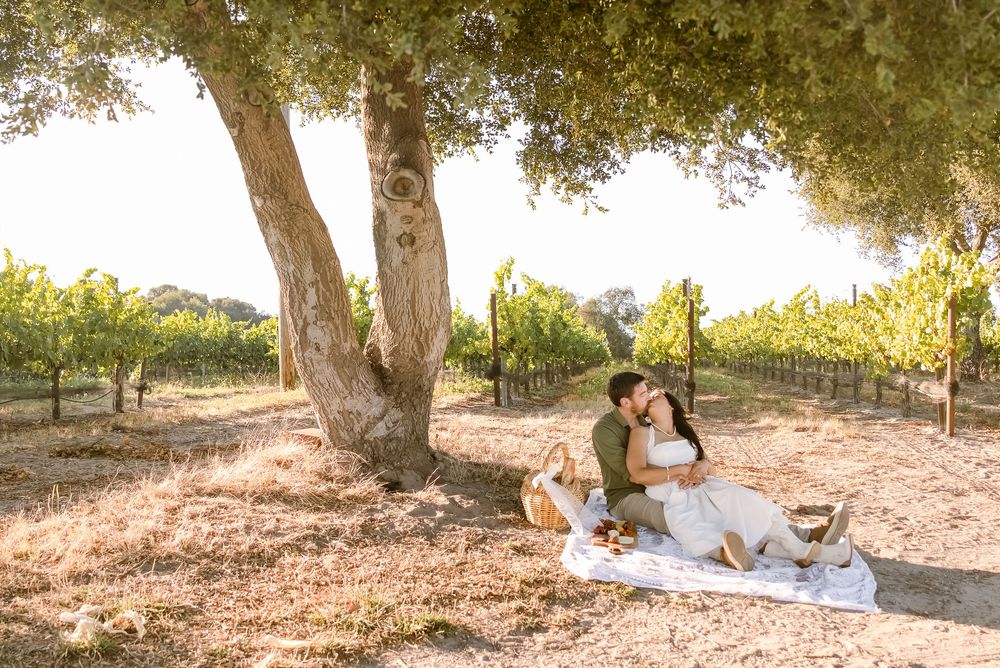 Couple sitting together under a large oak tree at a vineyard picnic, sharing a romantic outdoor moment.
