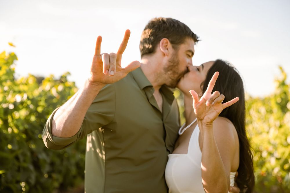 Couple kissing while showing off engagement rings in a sunny vineyard setting.
