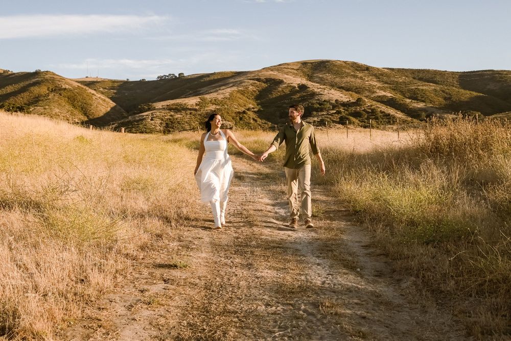Couple holding hands walking on a dirt path through golden hills landscape on a sunny day.