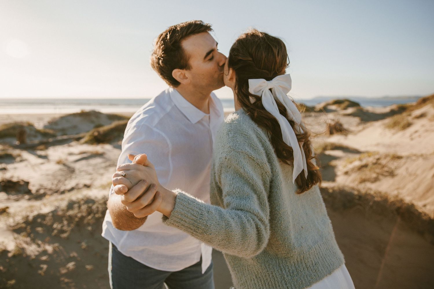 A couple shares a tender kiss on a sunny beach with sand dunes and blue sky in the background.