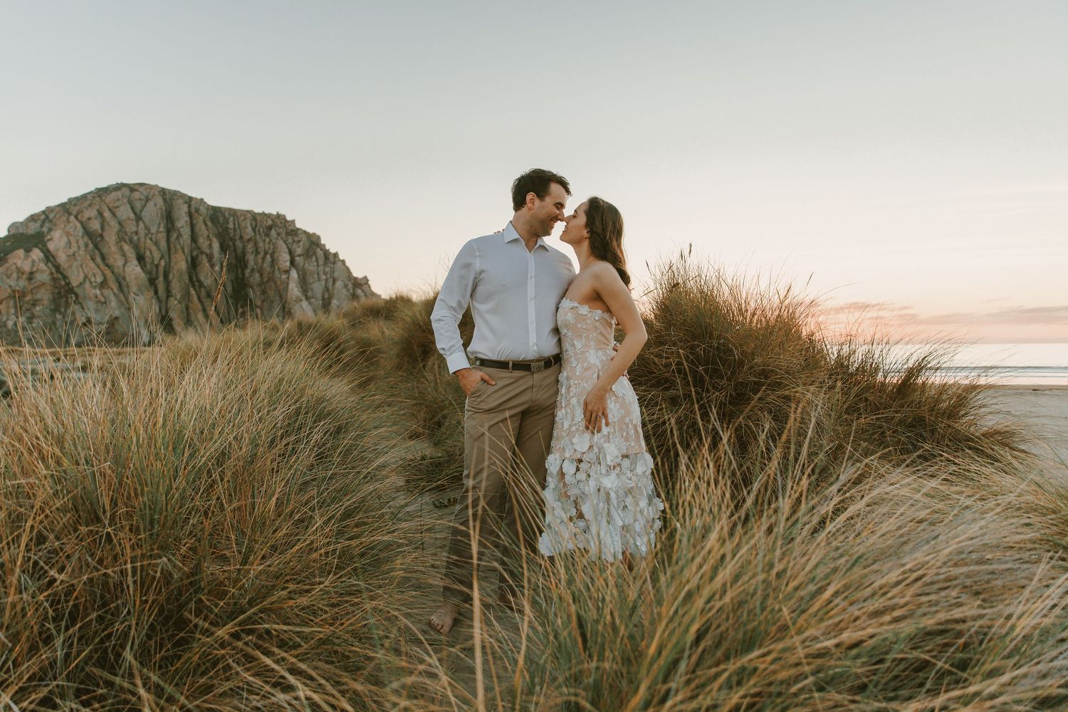 A couple stands close together in tall beach grass at sunset with Morro Rock visible in the background.