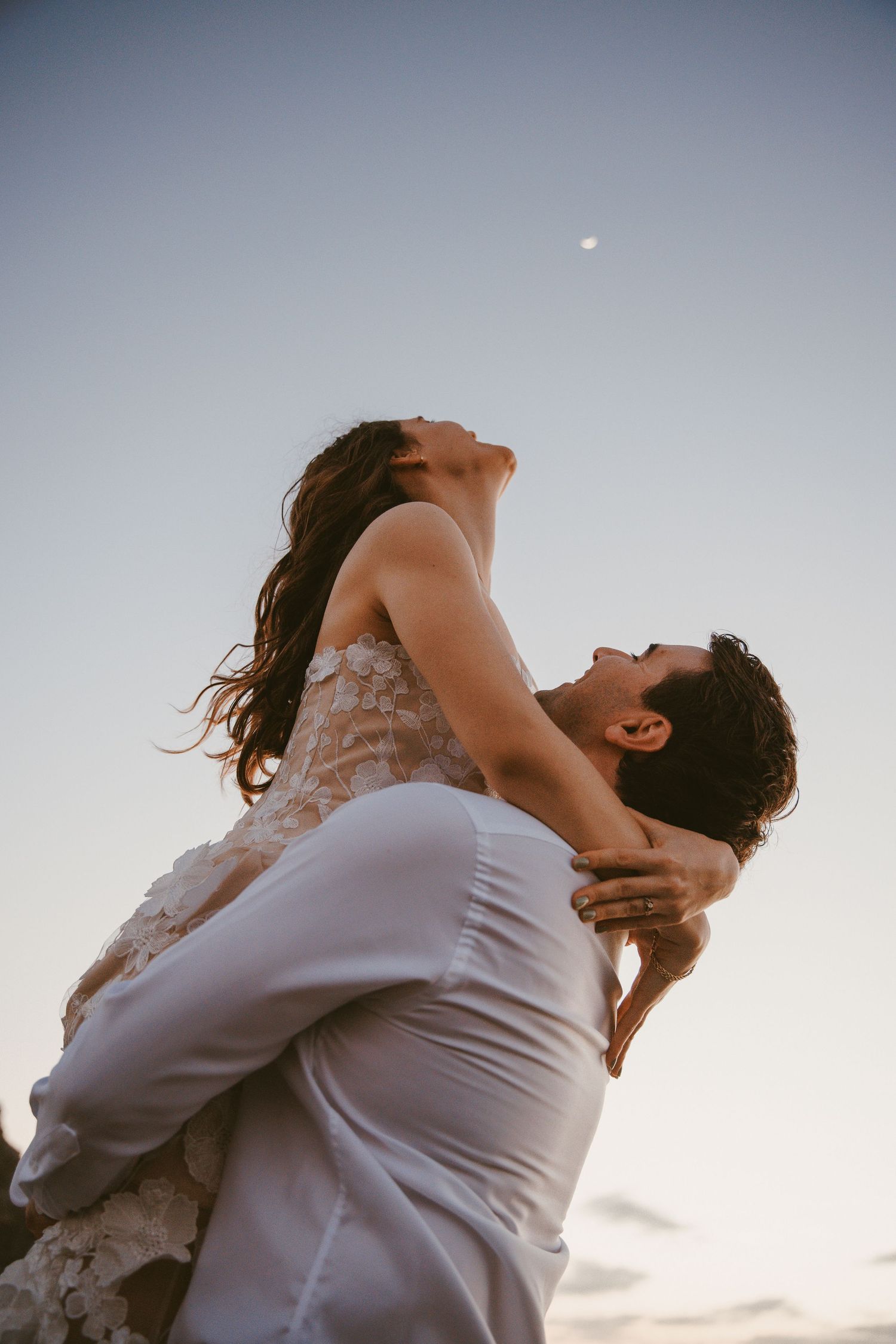 A man lifts a woman in a lace dress against a twilight sky with a faint moon visible above.