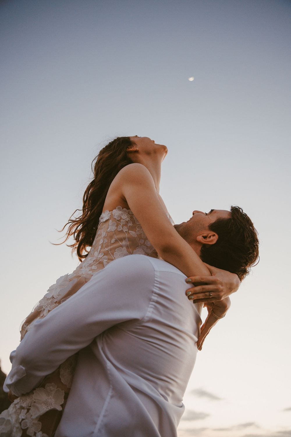 A man lifts a woman in a lace dress against a twilight sky with a faint moon visible above.