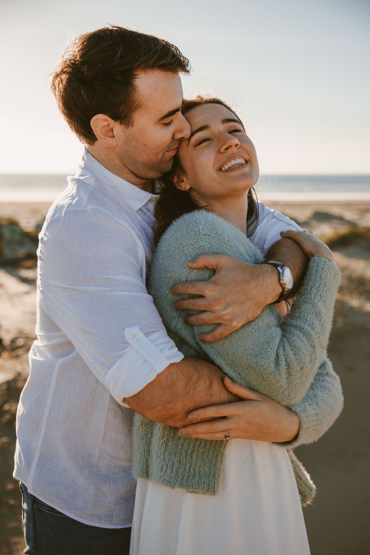 A man embraces a laughing woman from behind on a rocky beach at golden hour sunset.