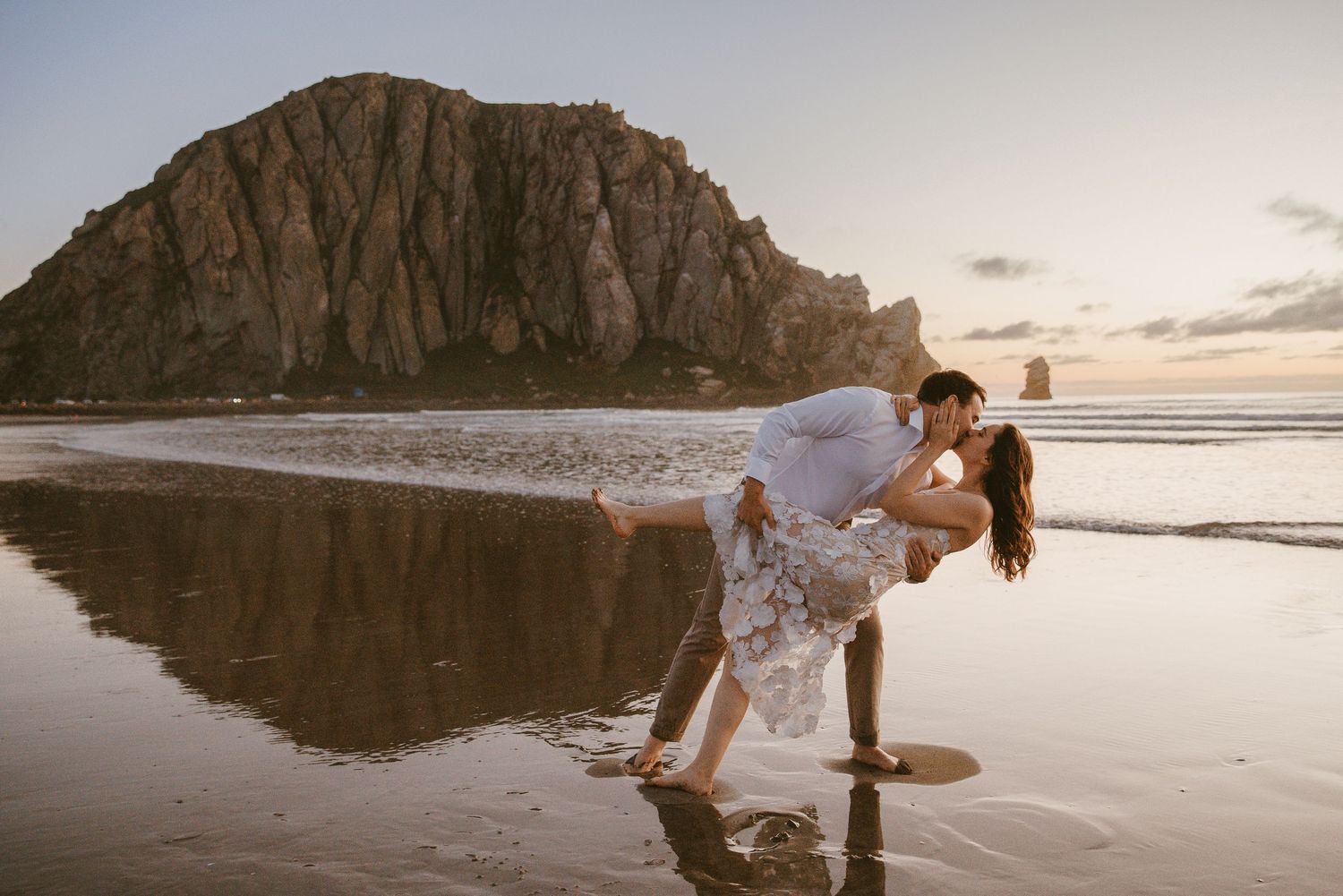 Romantic couple sharing a dip kiss on a beach at sunset with Morro Rock backdrop.
