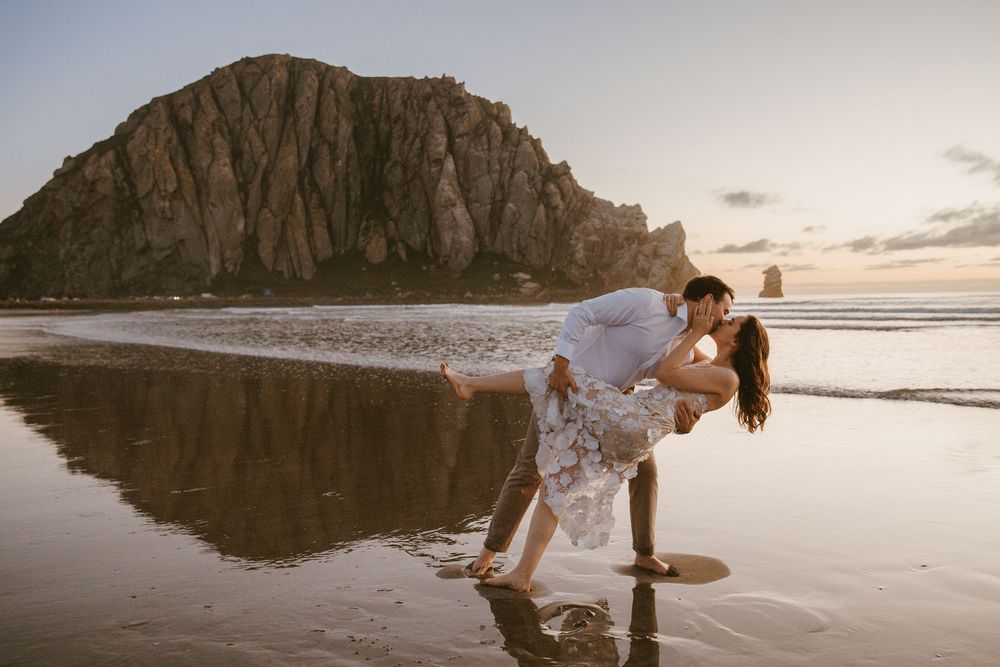 Romantic couple sharing a dip kiss on a beach at sunset with Morro Rock backdrop.