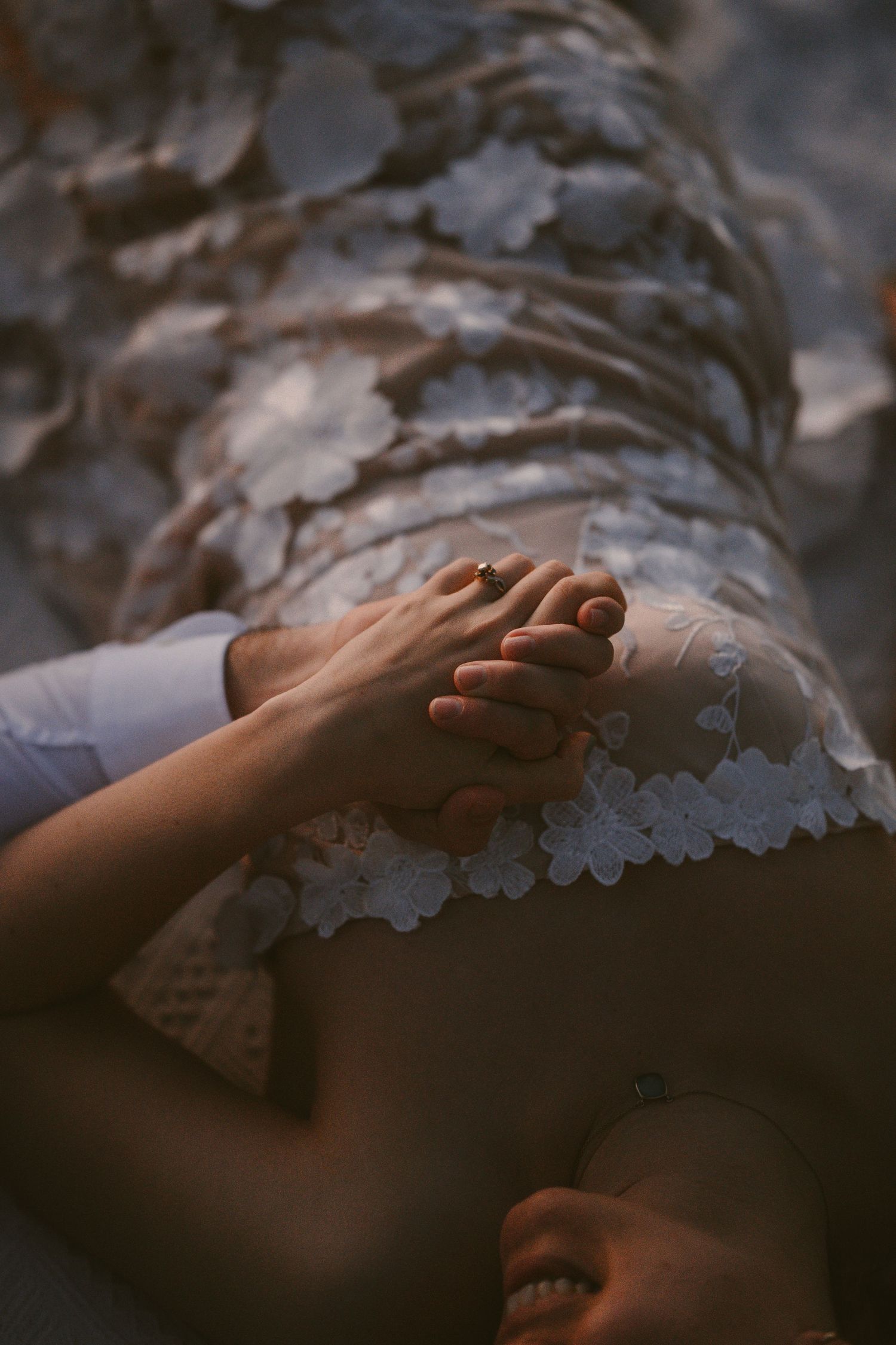 Couple holding hands tenderly over a floral lace wedding dress on rocky ground.