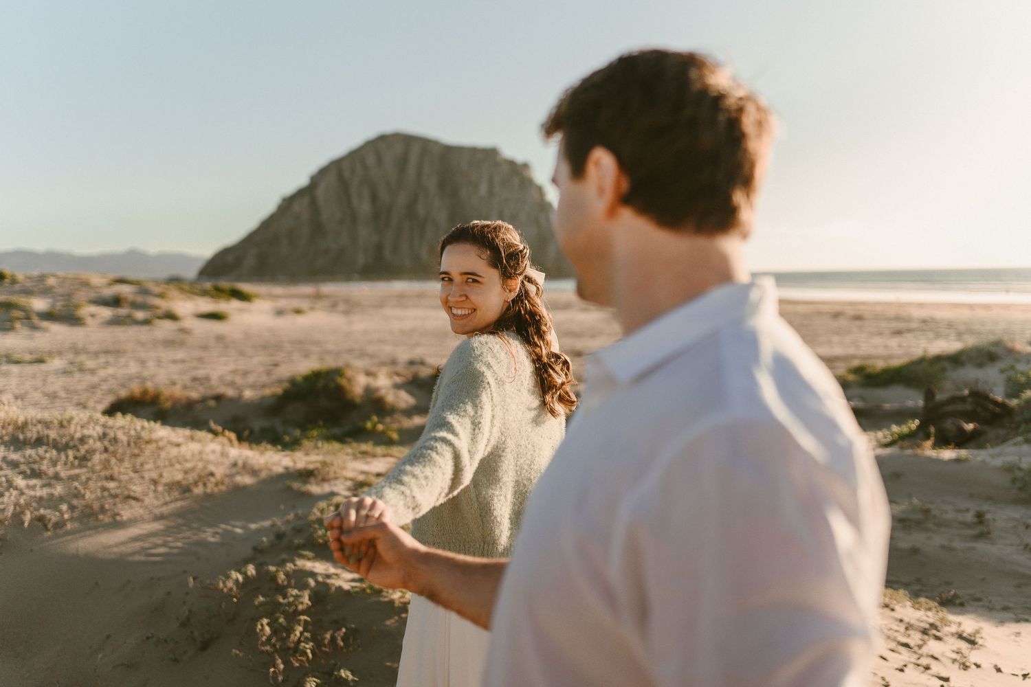 A smiling woman leads her partner by the hand along a coastal beach path with Morro Rock in the distance.