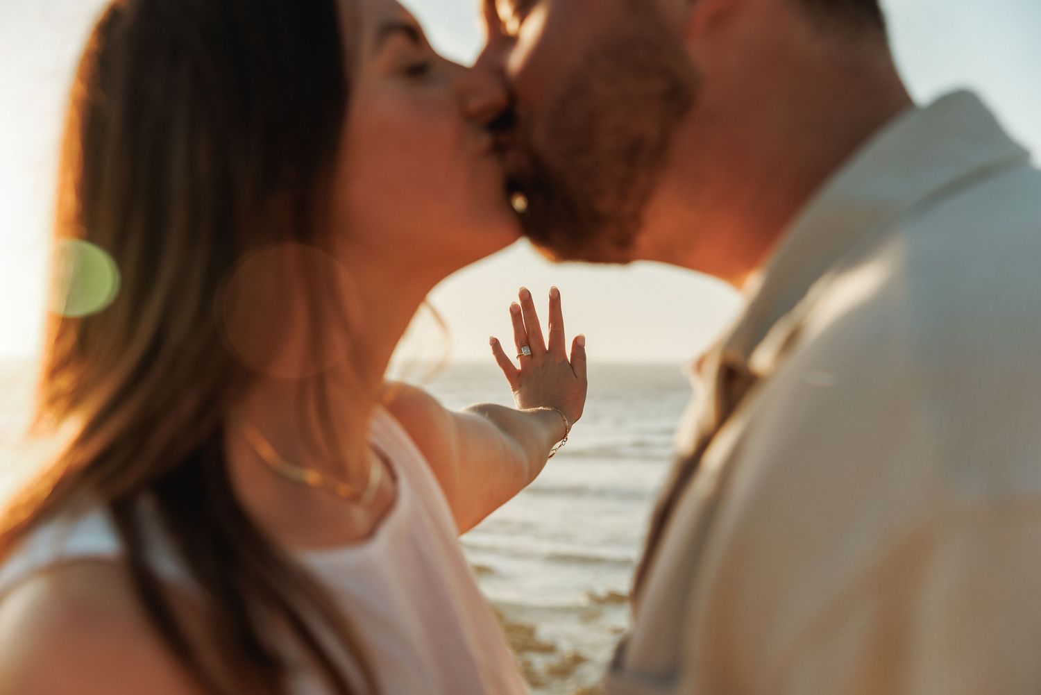 Close-up of couple kissing at beach sunset, woman displaying engagement ring toward camera.