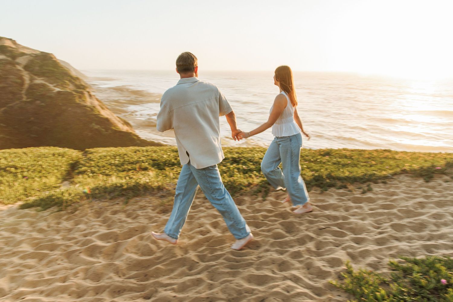 Couple holding hands walking on sandy path toward ocean at golden hour sunset.