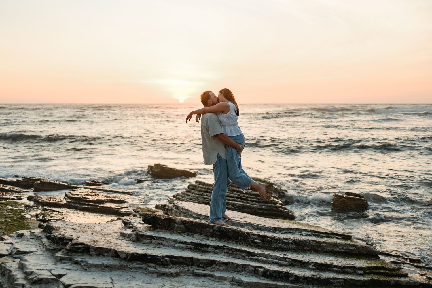 Couple embracing on rocky shoreline with waves and pink sunset sky in background.