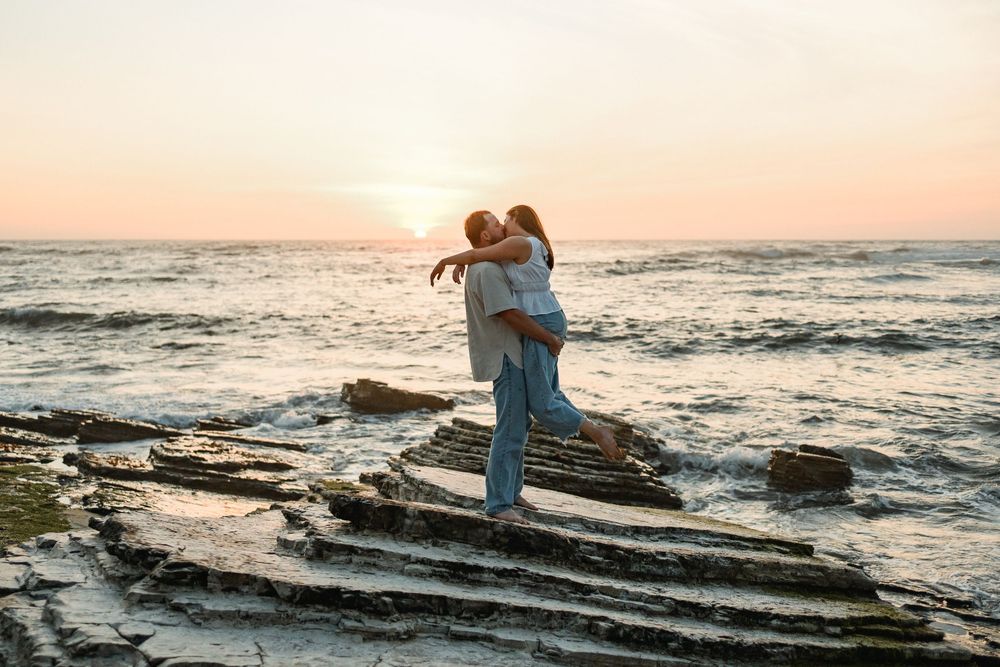 Couple embracing on rocky shoreline with waves and pink sunset sky in background.