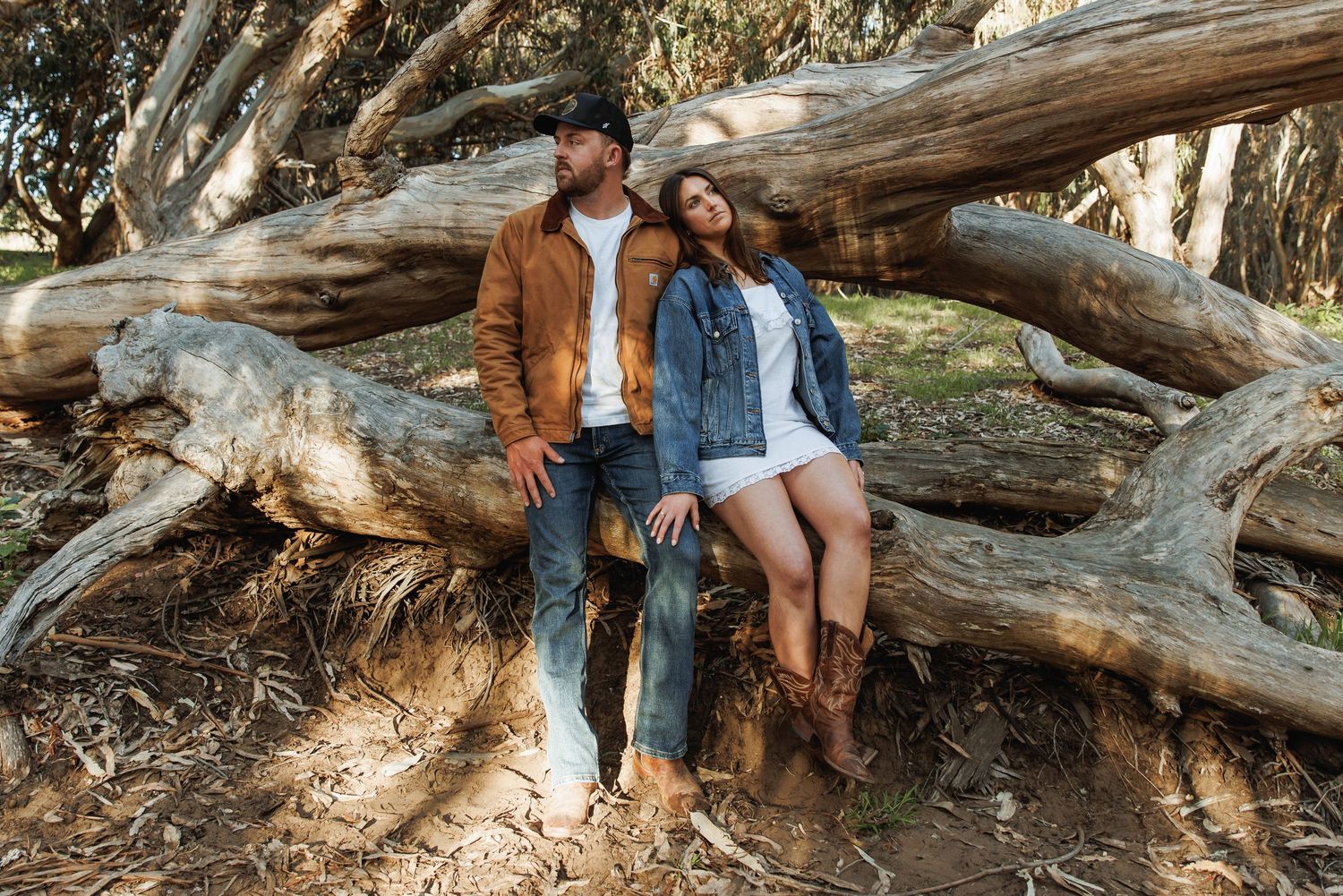 Couple leaning against large tree roots in a forest, man in brown jacket and woman in denim jacket.